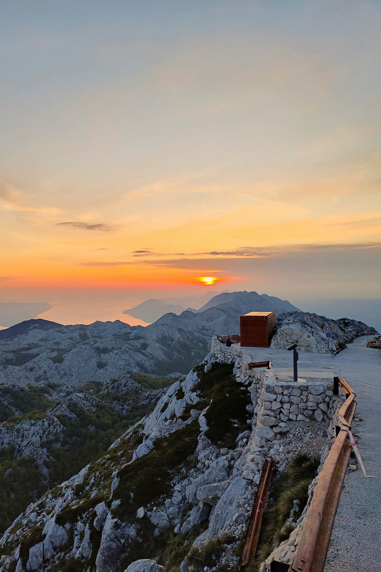 Bild av en bergstopp under solnedgång med stenmur, en gångväg och ett räcke i förgrunden. (AI-genererad text)
