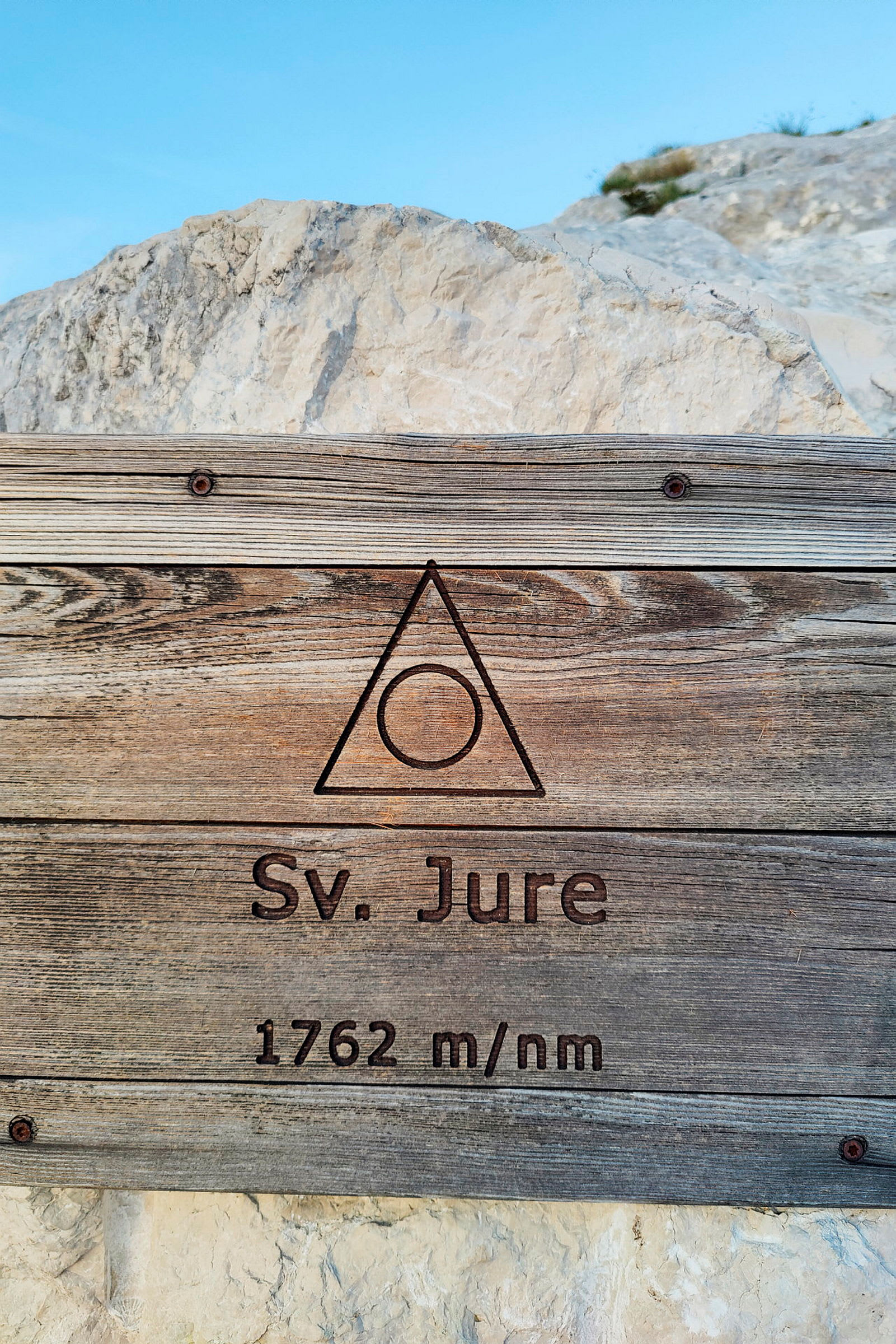Träskylt med texten 'Sv. Jure 1762 m/nm' mot en bakgrund av klippor och blå himmel. (AI-genererad text)