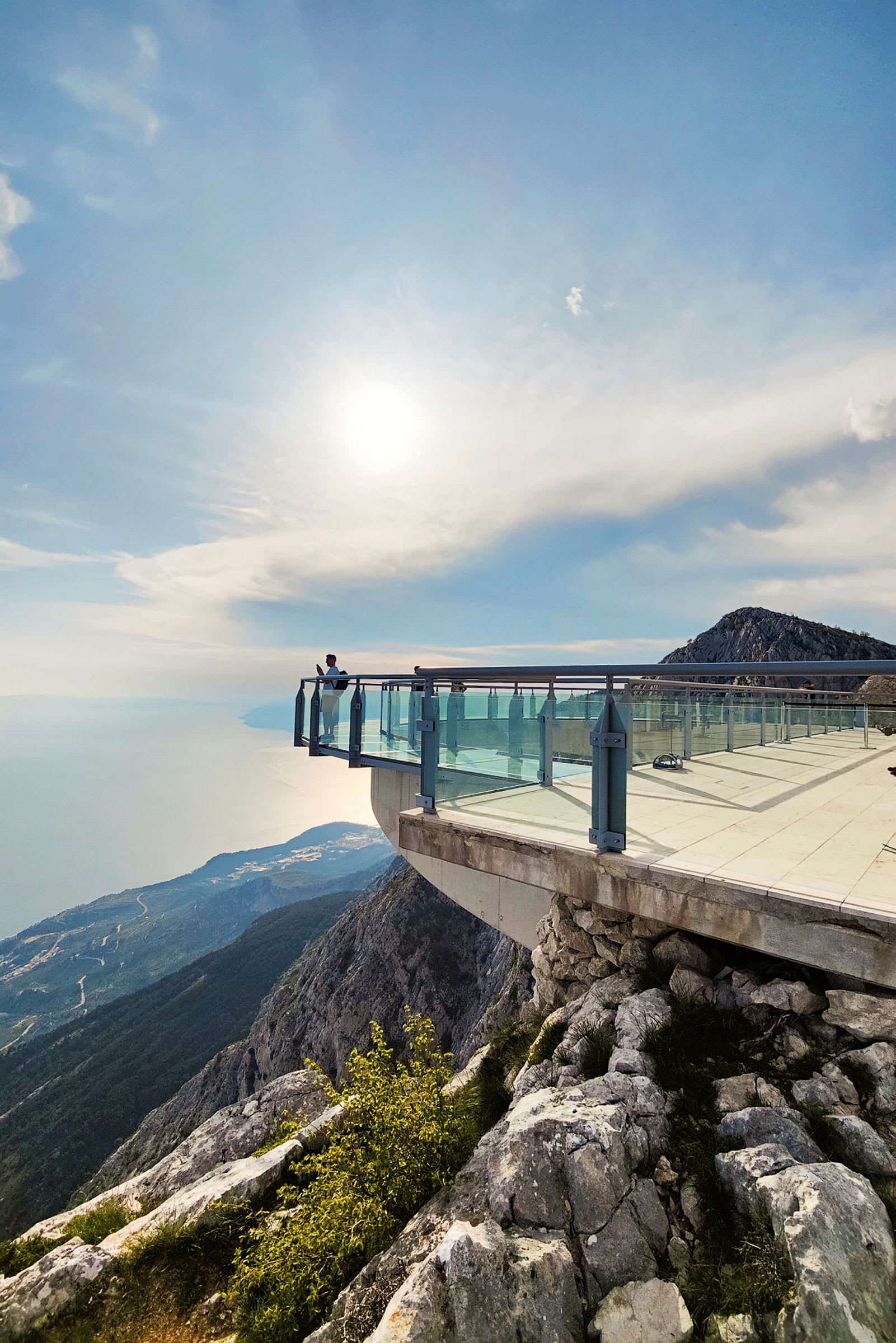 En utsiktspunkt med glasräcke som sträcker sig ut över en klippa. Solen lyser och skapar ett mjukt ljus över bergslandskapet och havet nedanför. (AI-genererad text)