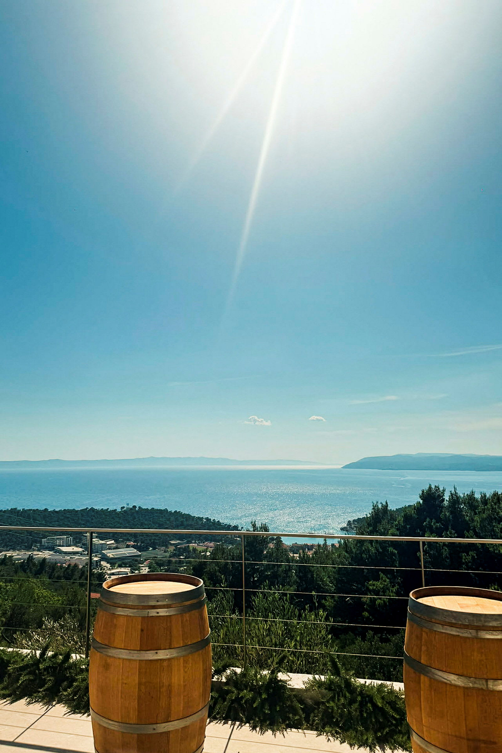 Två träfat står på en terrass med utsikt över ett glittrande hav och grönskande landskap under en klar himmel. (AI-genererad text)