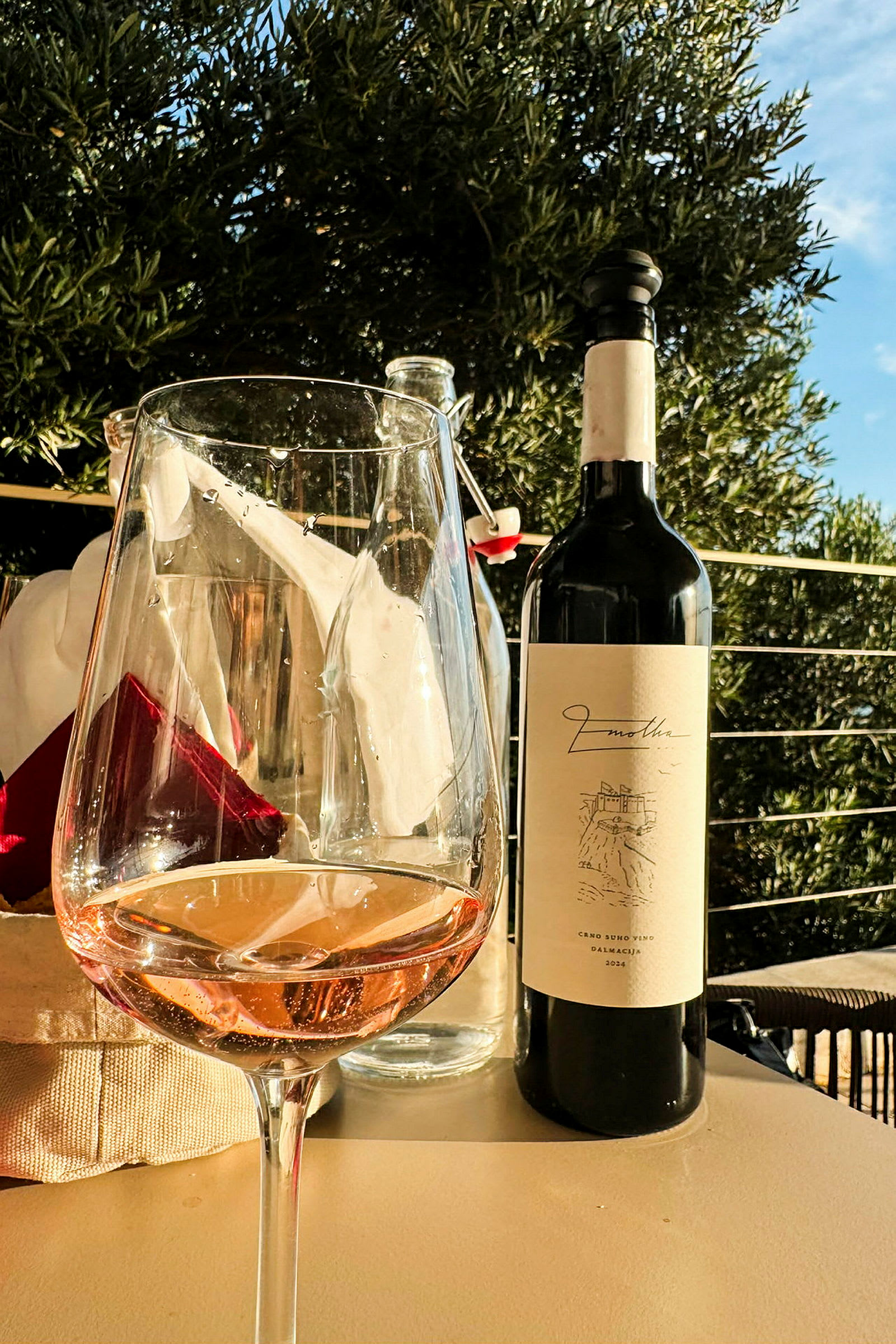 Ett glas rosévin och en vinflaska står på ett bord i en utomhusmiljö med grönska i bakgrunden. (AI-genererad text)