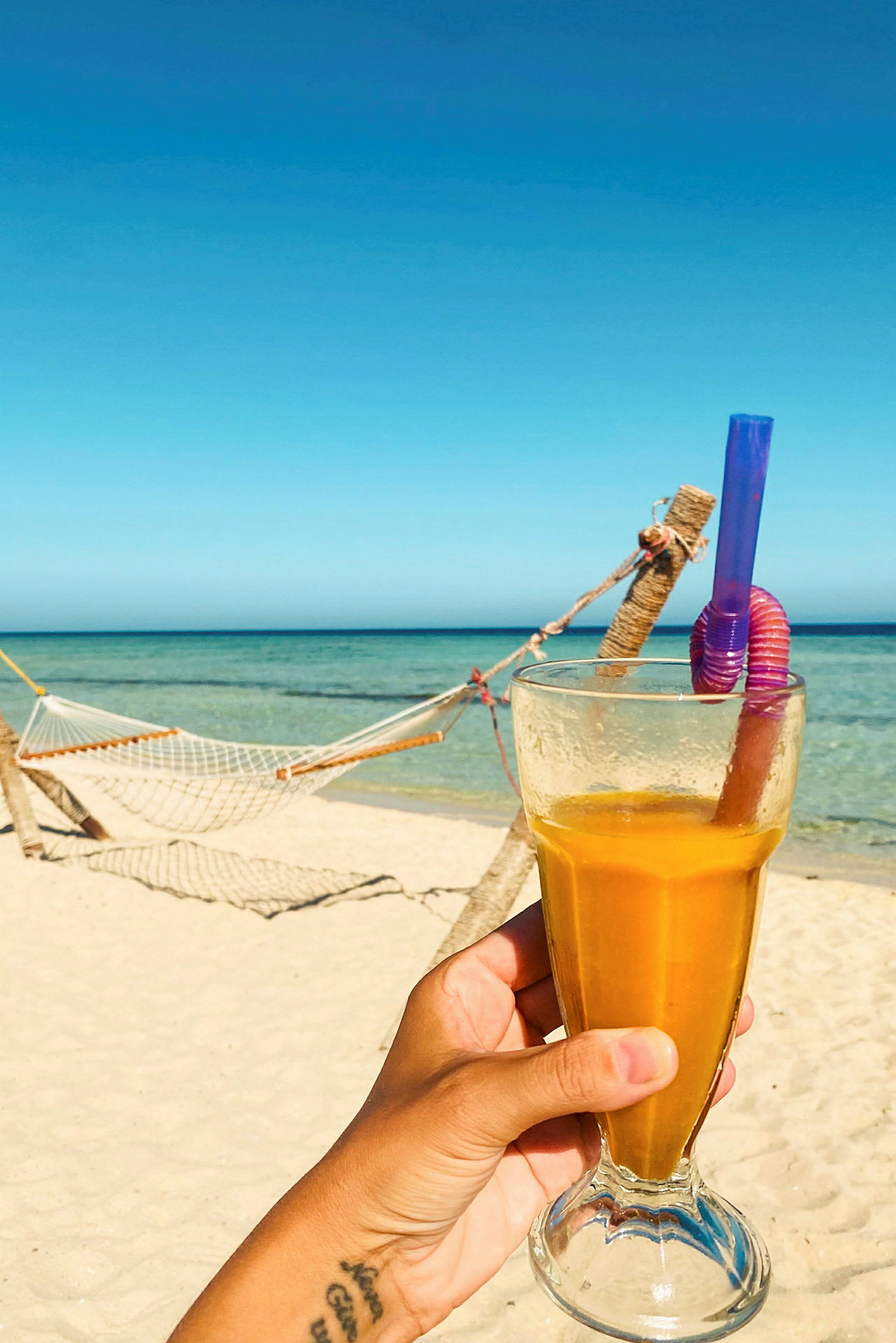 Ett glas med gul dryck hålls av en hand på en strand med en hängmatta och hav i bakgrunden. (AI-genererad text)