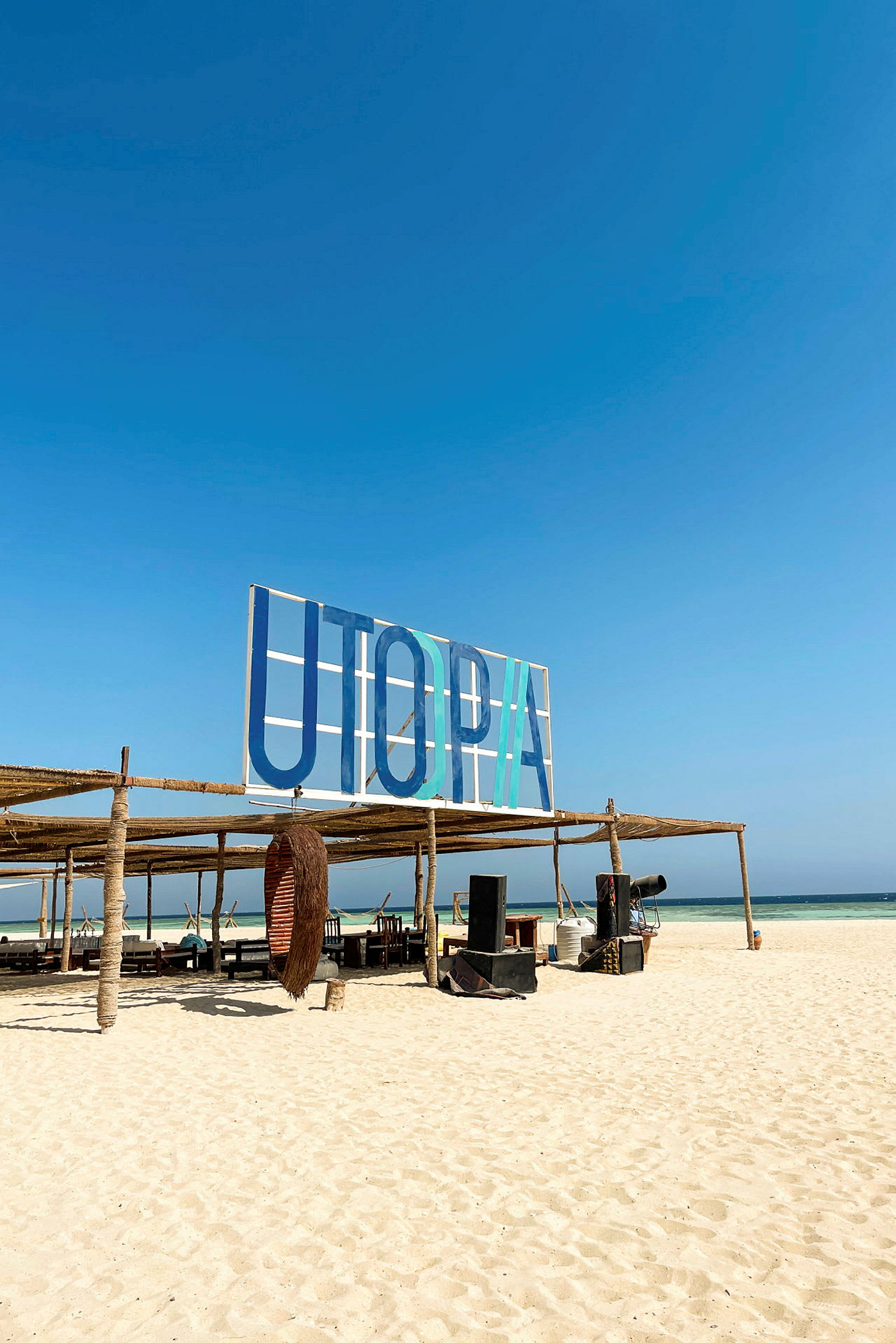 Skylt med texten 'UTOPIA' på en strand med sittplatser och rekvisita. (AI-genererad text)