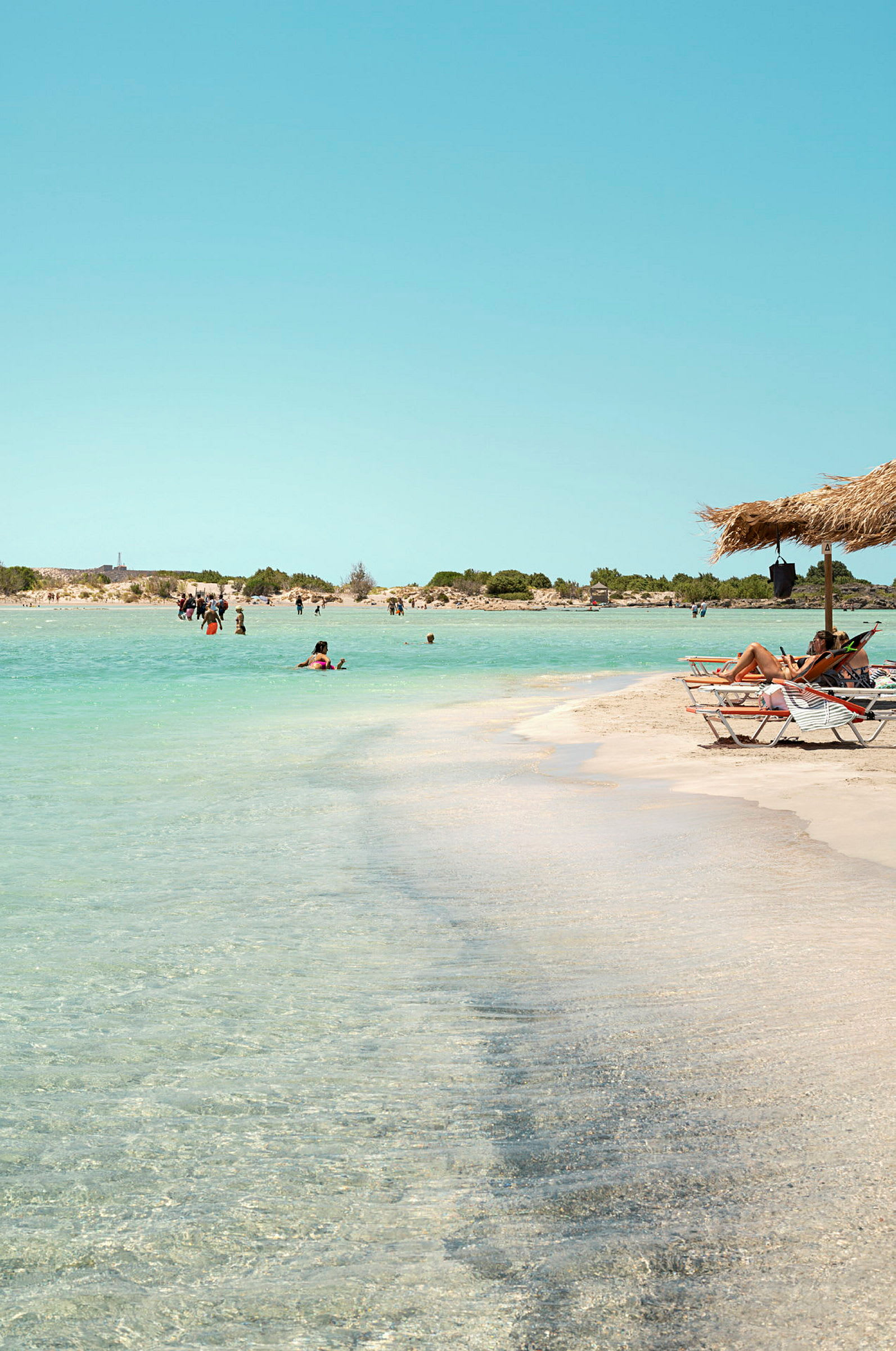 En strand med vit sand och klart vatten, omgiven av låga buskar. Flera personer badar i havet och några sitter i solstolar under ett parasoll. (AI-genererad text)