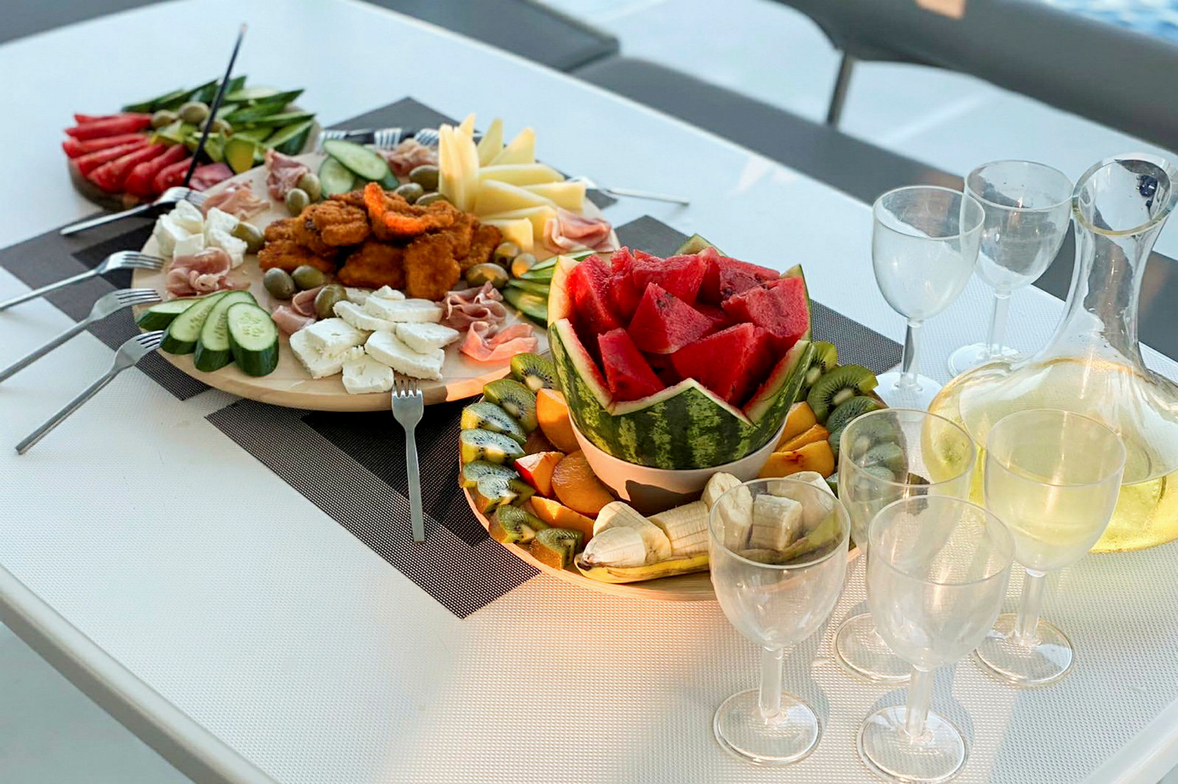 En bild av ett dukat bord med ett fat frukt, inklusive vattenmelon och kiwifrukt, samt en skål med sallad och kött. Glas och en karaff med dryck finns på bordet. (AI-genererad text)