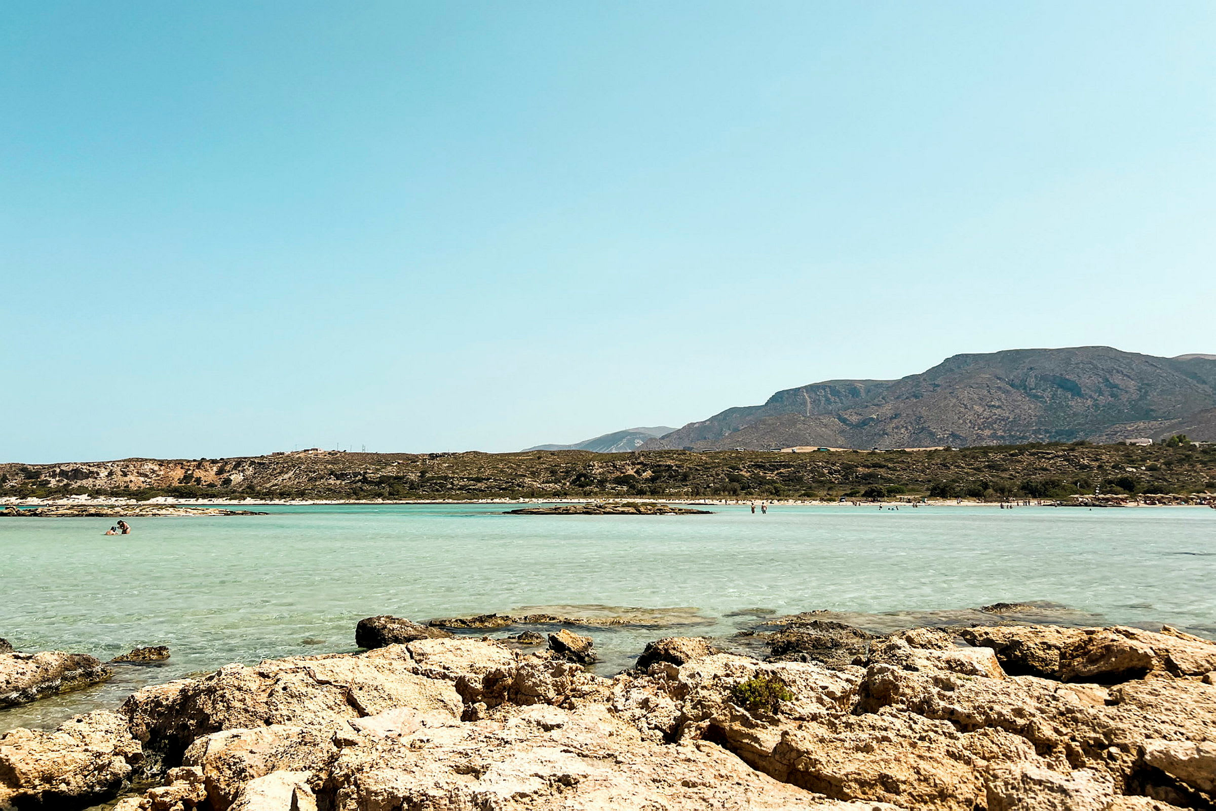 En strand med klippor i förgrunden, klart blått vatten och berg i bakgrunden. (AI-genererad text)