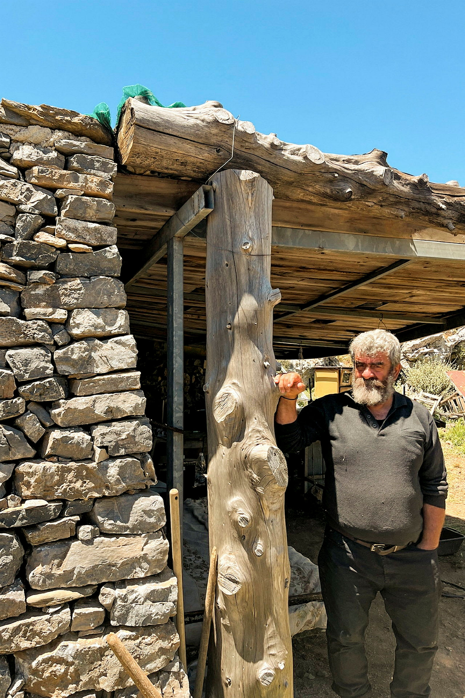 En person står bredvid en träpelare och en vägg av natursten under en enkel trästruktur utomhus. (AI-genererad text)