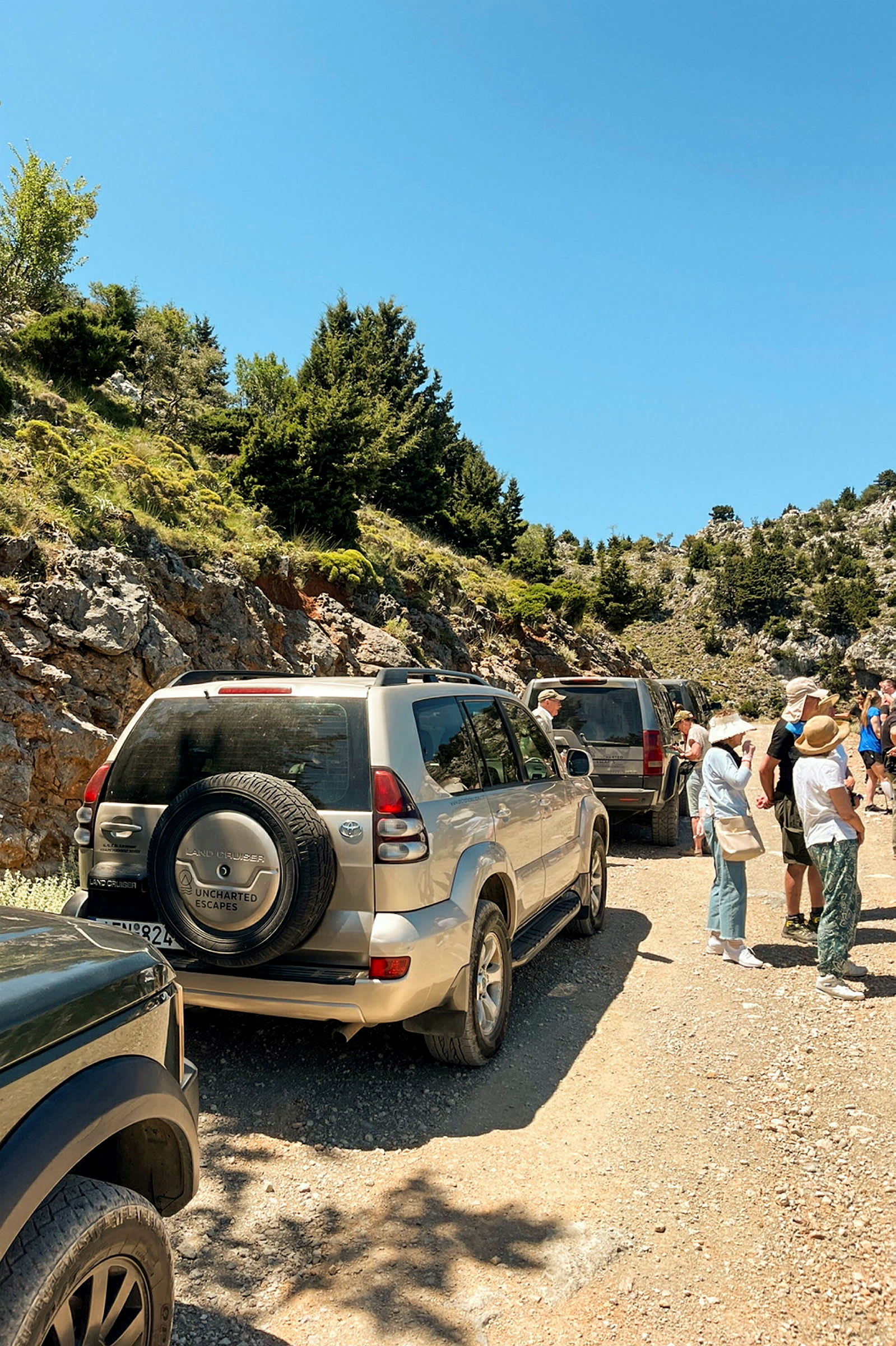 En grupp terrängbilar står parkerade vid en grusväg omgiven av bergig och grönskande natur, där människor samlas vid sidan av vägen. (AI-genererad text)