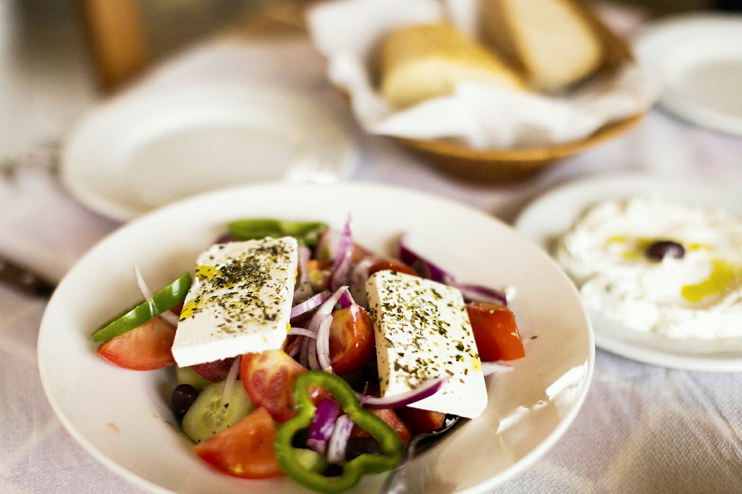 En tallrik med grekisk sallad innehållande tomater, gurka, paprika, lök och fetaost. I bakgrunden syns bröd och en tallrik med yoghurt och olivolja. (AI-genererad text)