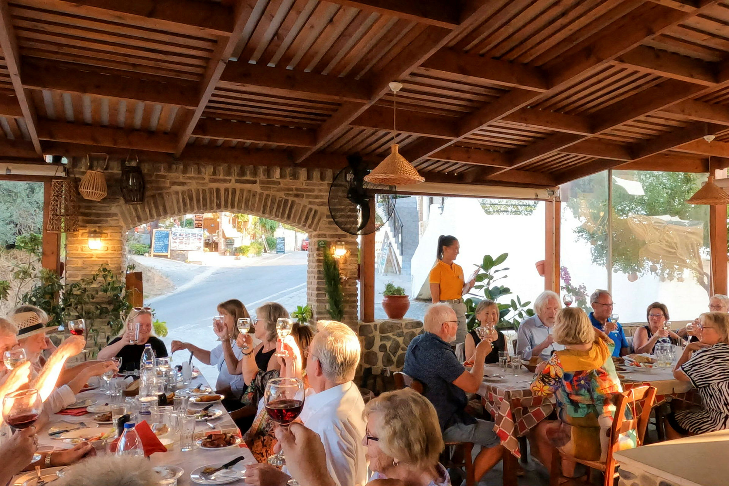 En grupp personer sitter runt ett långbord på en utomhusrestaurang under ett träpergola. (AI-genererad text)