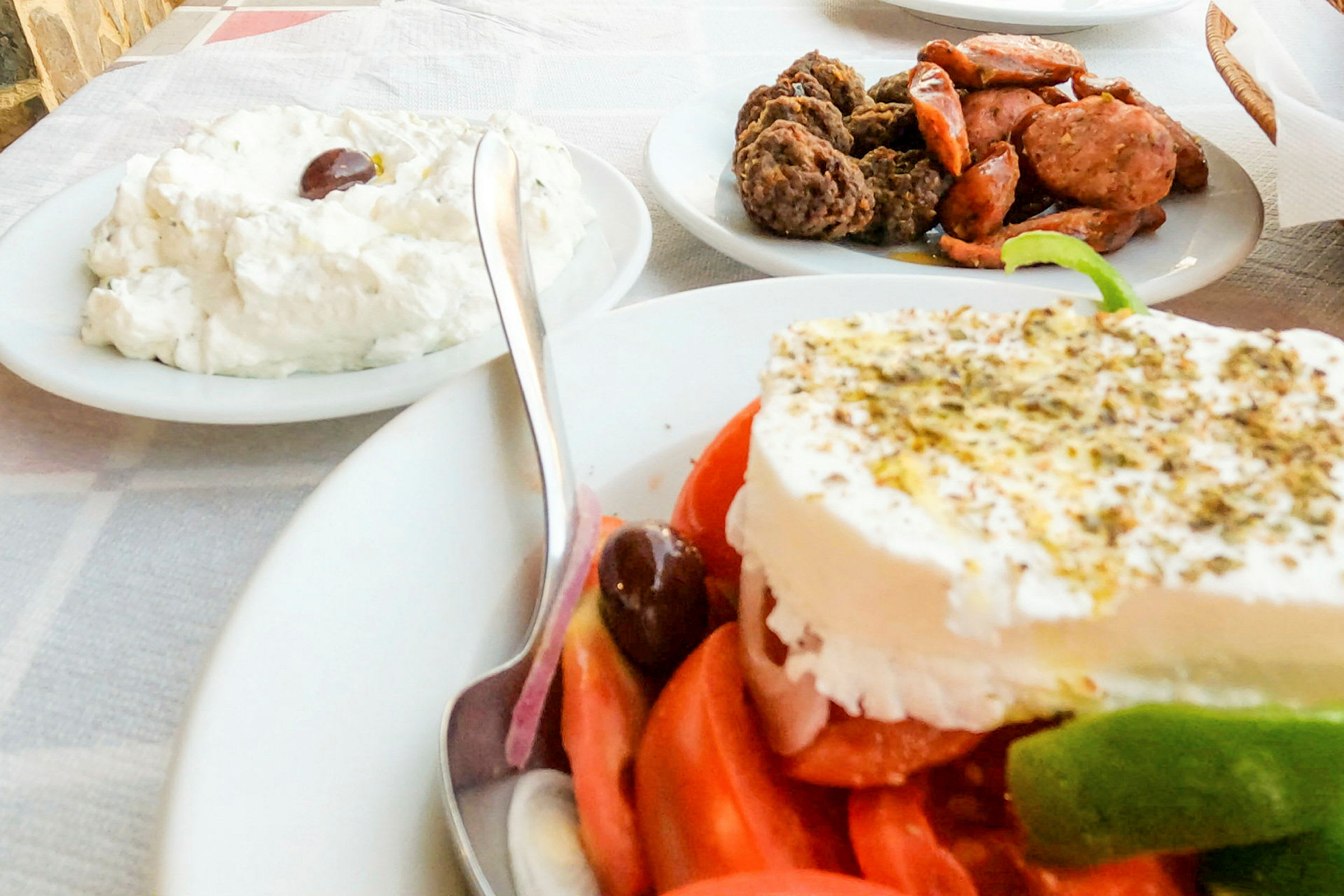 En tallrik med en grekisk sallad toppad med fetaost, en skål med tzatziki och en tallrik med kött och korv på ett bord. (AI-genererad text)