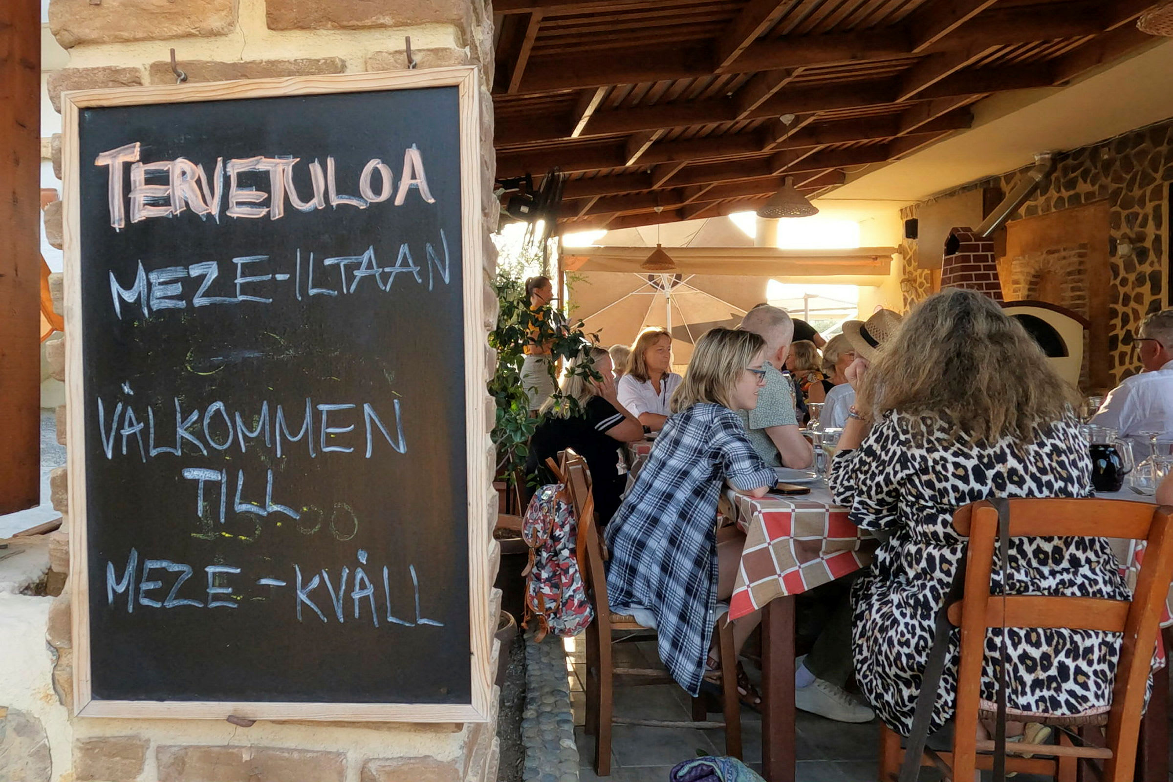 En svart tavla med texten "Välkommen till meze-kväll" står vid ingången till en restaurang med gäster sittande vid bord. (AI-genererad text)