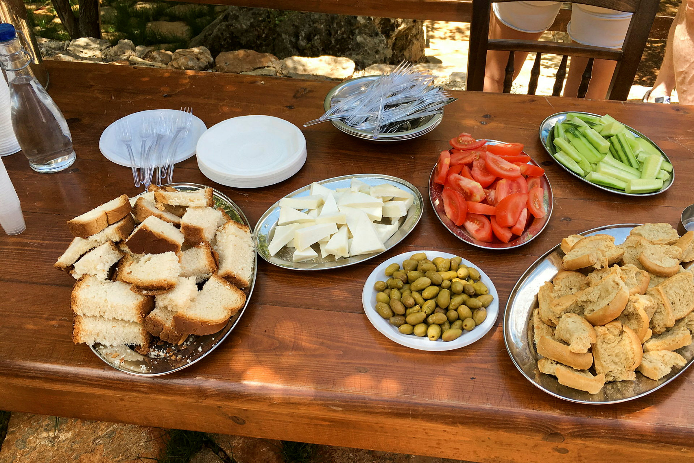 Ett bord med olika matvaror, inklusive bröd, ost, oliver, tomater och gurkor. Det finns också tallrikar, bestick och en vattenflaska. (AI-genererad text)