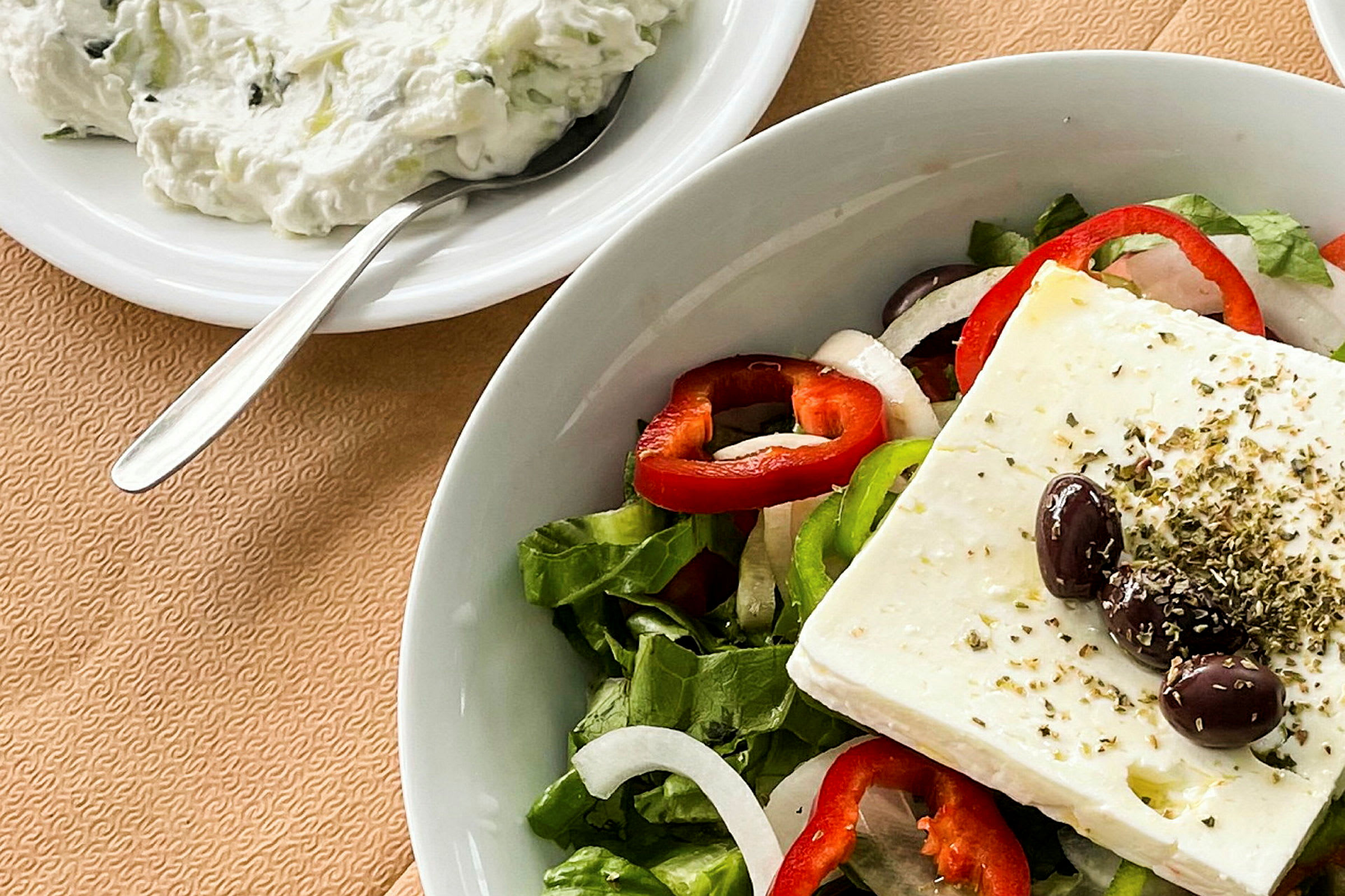 En tallrik med en sallad bestående av fetaost, oliver, paprikaringar, lök och salladsblad. Bredvid står en skål med tzatziki och en sked. (AI-genererad text)