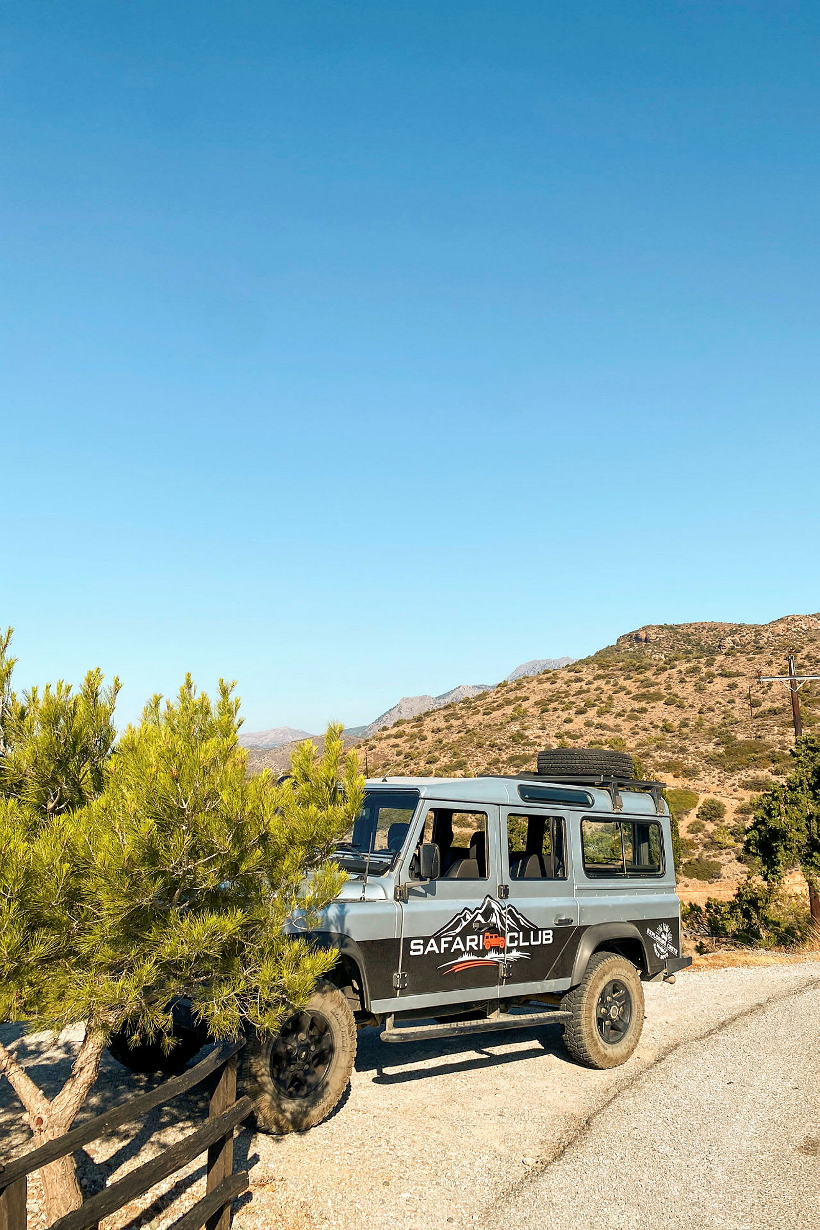En fyrhjulsdriven jeep med texten "Safari Club" på sidan står parkerad på en grusväg bredvid ett träd, med berg och klar himmel i bakgrunden. (AI-genererad text)