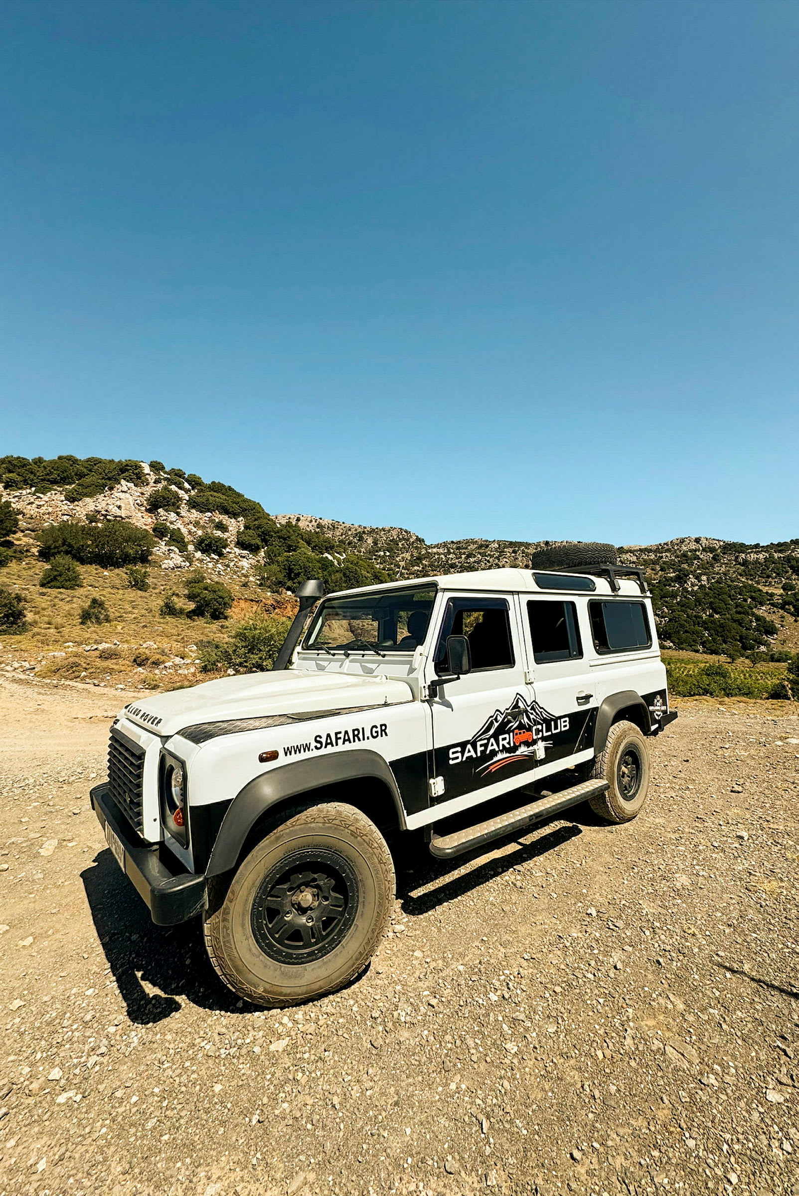 En vit fyrhjulsdriven bil med texten "Safari Club" står parkerad på en grusväg i ett bergigt landskap. (AI-genererad text)