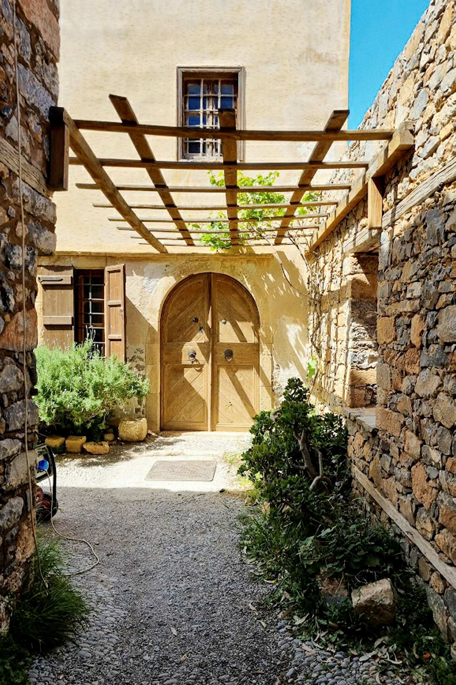 En gammal byggnad med en trädörr och stenmur omgiven av växter under en pergola. (AI-genererad text)