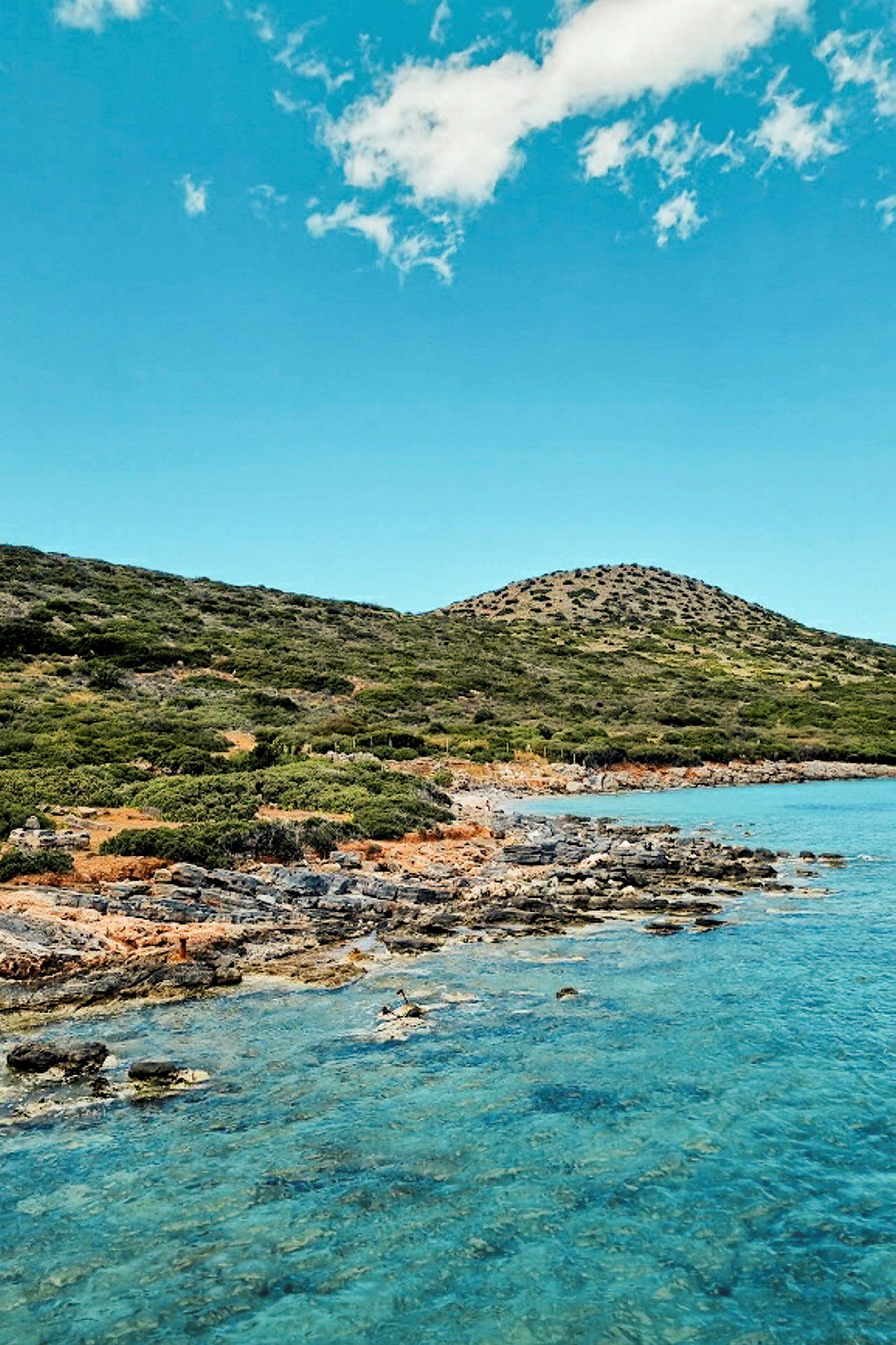 Strandlinje med klippor och buskig vegetation, samt klart turkost havsvatten under en blå himmel. (AI-genererad text)