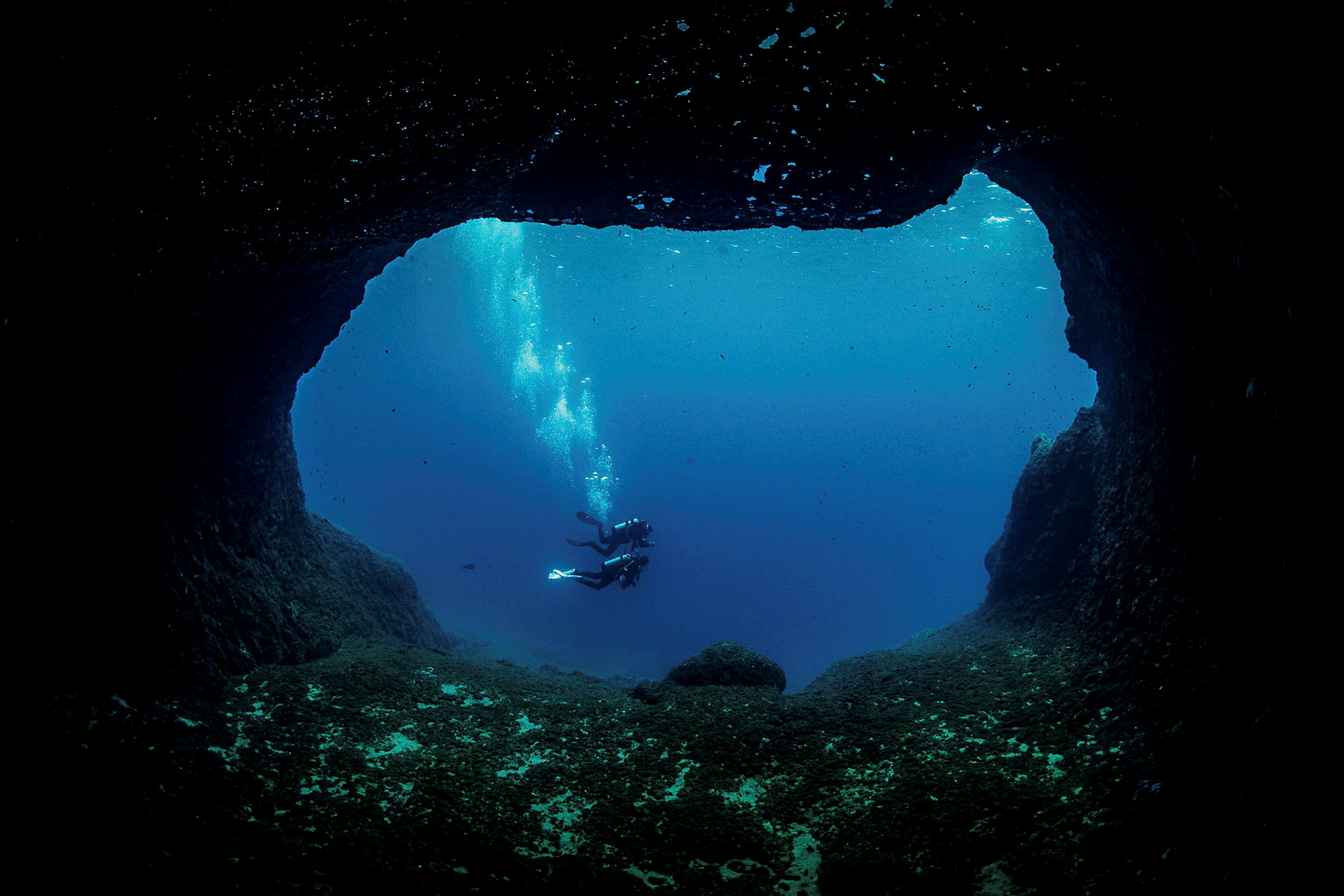 Två dykare simmar i havet, sett från insidan av en undervattensgrotta. (AI-genererad text)