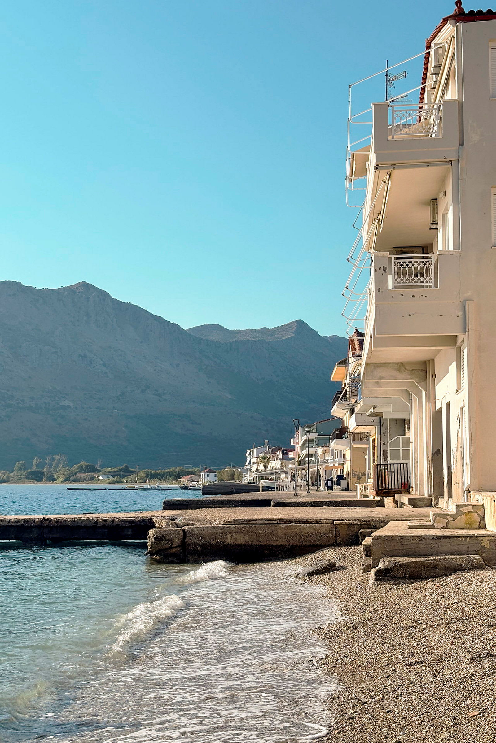 Vy över strandpromenad med byggnader vid stranden och berg i bakgrunden. (AI-genererad text)