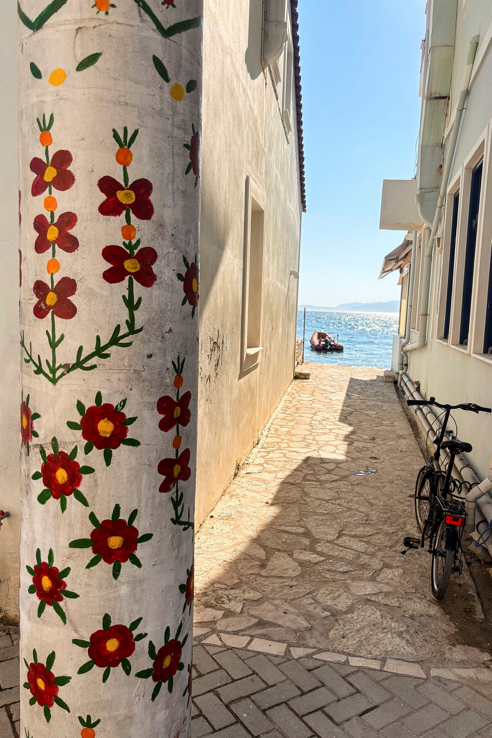 En smal stenig passage mellan hus, med en pelare dekorerad med målade röda blommor i förgrunden och havet synligt i bakgrunden. (AI-genererad text)