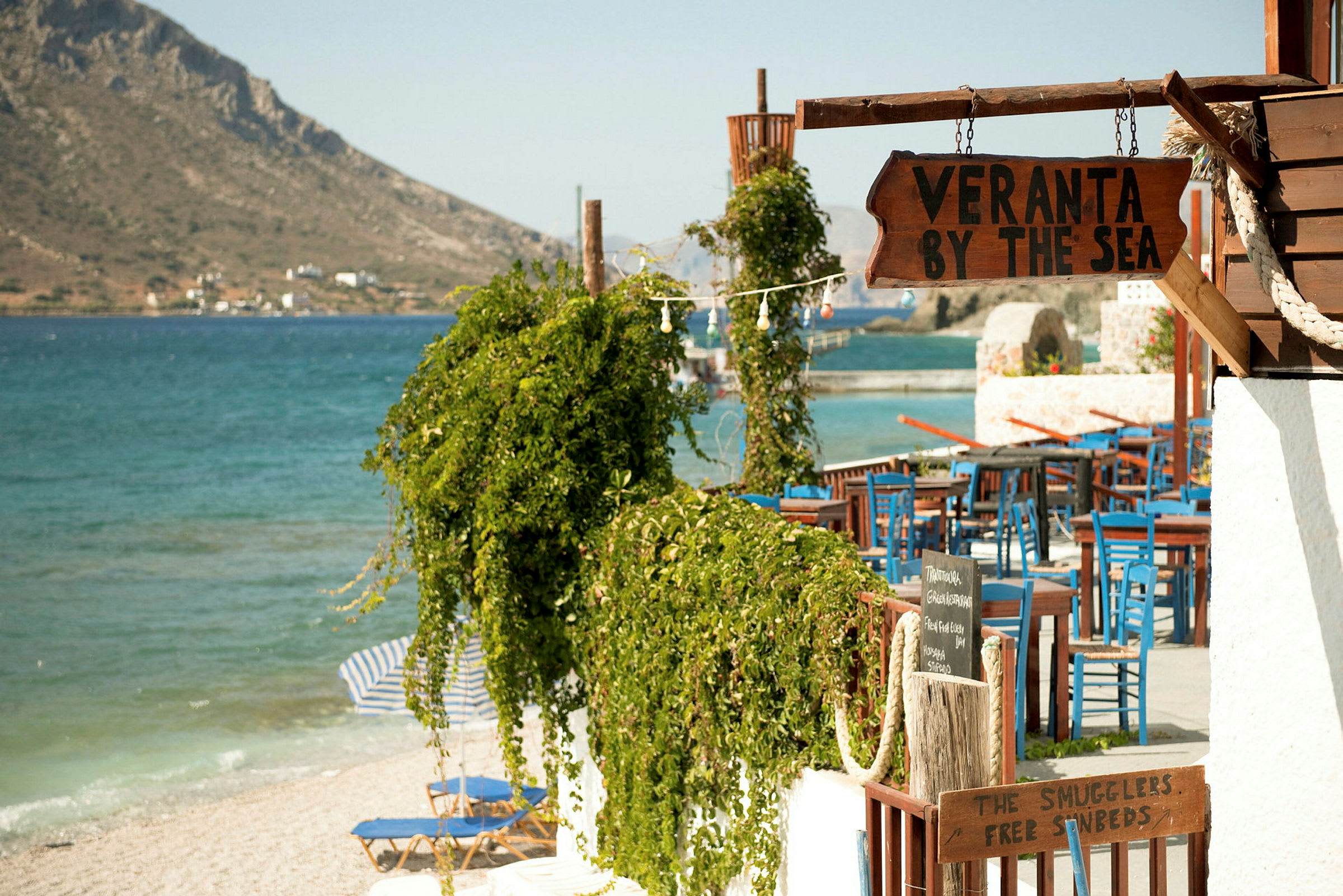 En närbild av en restaurang eller veranda vid stranden med skyltar som visar "Veranta by the sea" och blå stolar samt ett grönområde i förgrunden. (AI-genererad text)