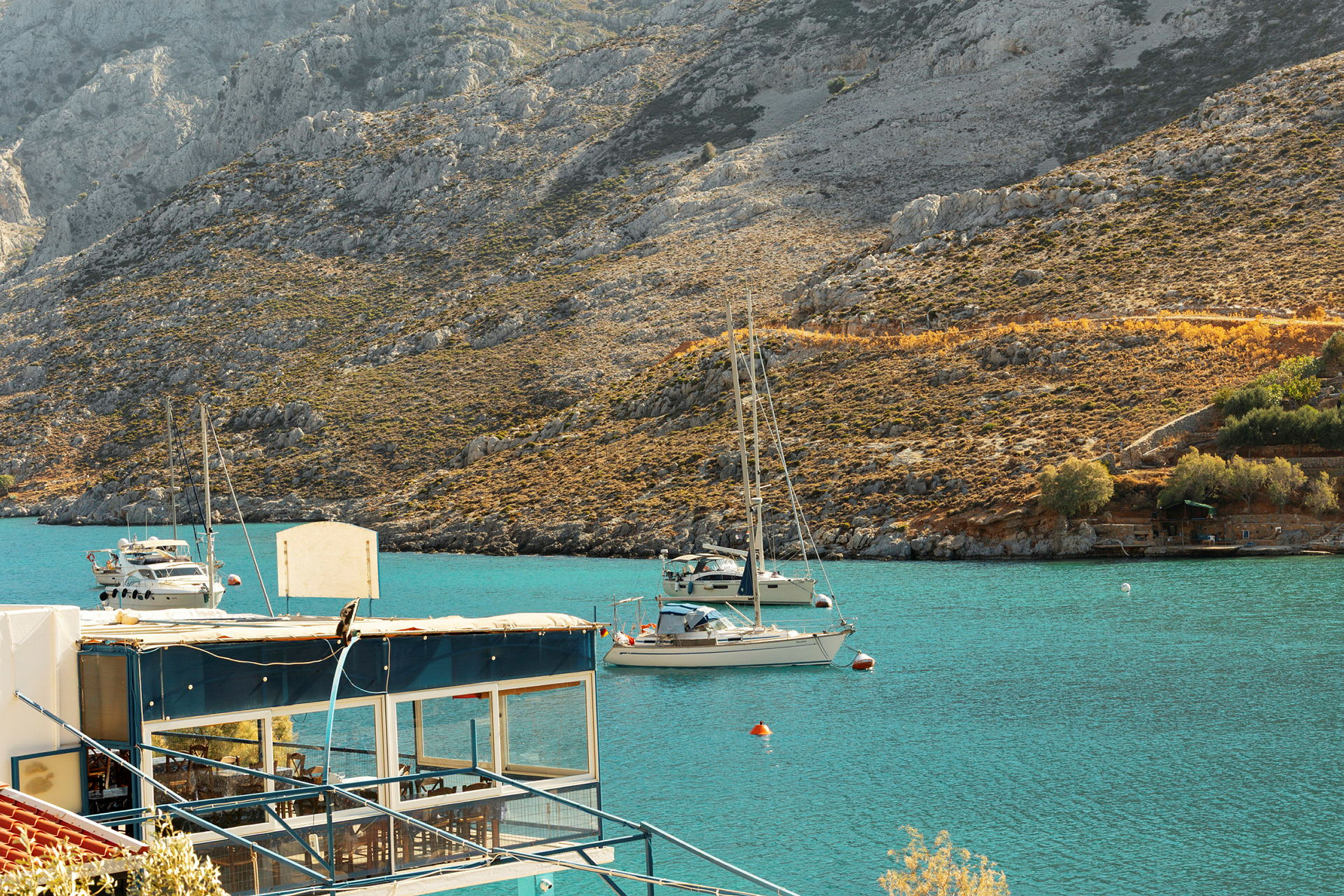 En vy över en bukt med klart blått vatten, segelbåtar och berg i bakgrunden. Ett hus med blåa detaljer syns i förgrunden. (AI-genererad text)