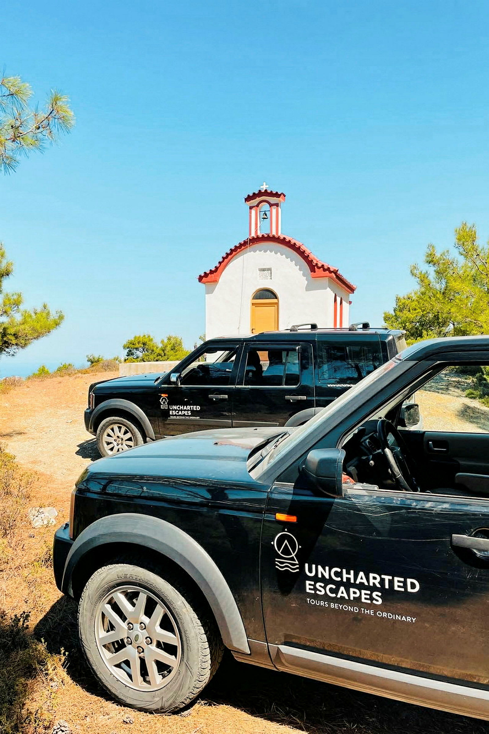 Två svarta bilar med logotypen "Uncharted Escapes" står parkerade framför en liten, vit kyrka med rött tak. Kyrkan är omgiven av grönska och himlen är klarblå. (AI-genererad text)