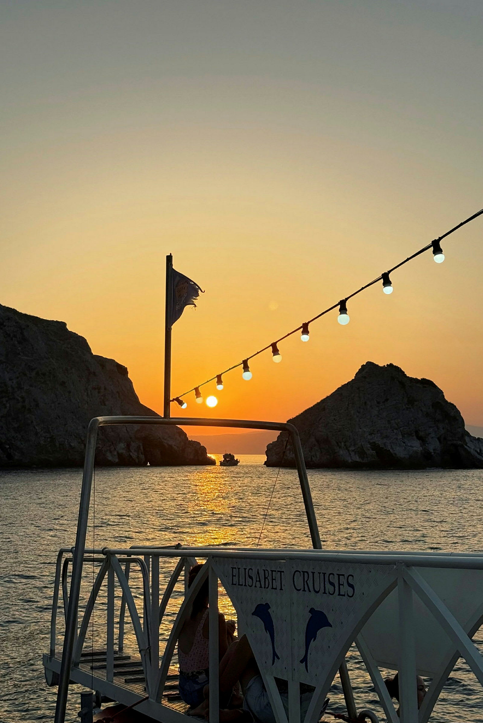 En solnedgång över havet med en brygga i förgrunden som bär en skylt med texten 'Elisabet Cruises'. I bakgrunden syns klippor och en båt. (AI-genererad text)