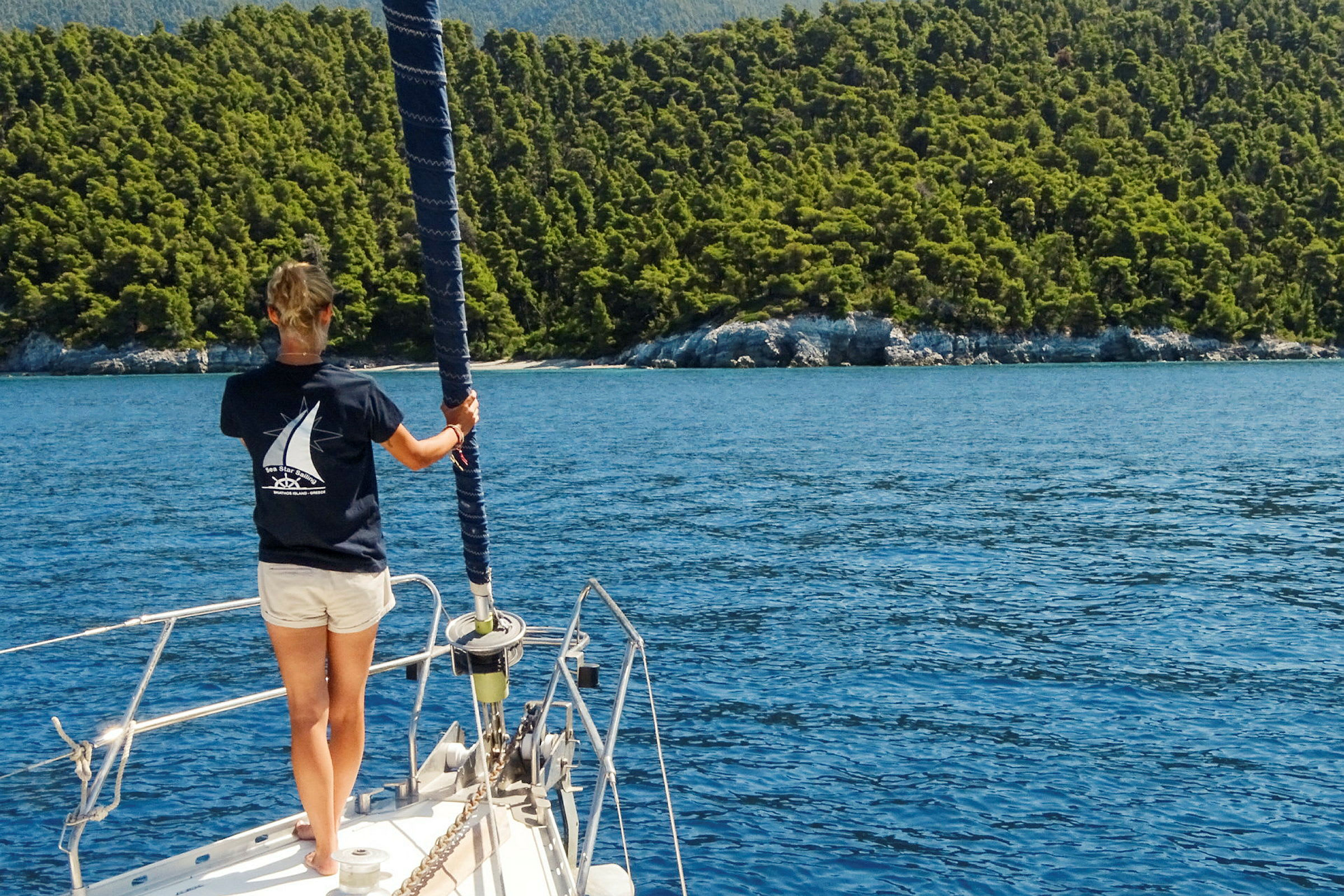En person står på däck på en segelbåt och tittar mot en skogbevuxen kust. (AI-genererad text)