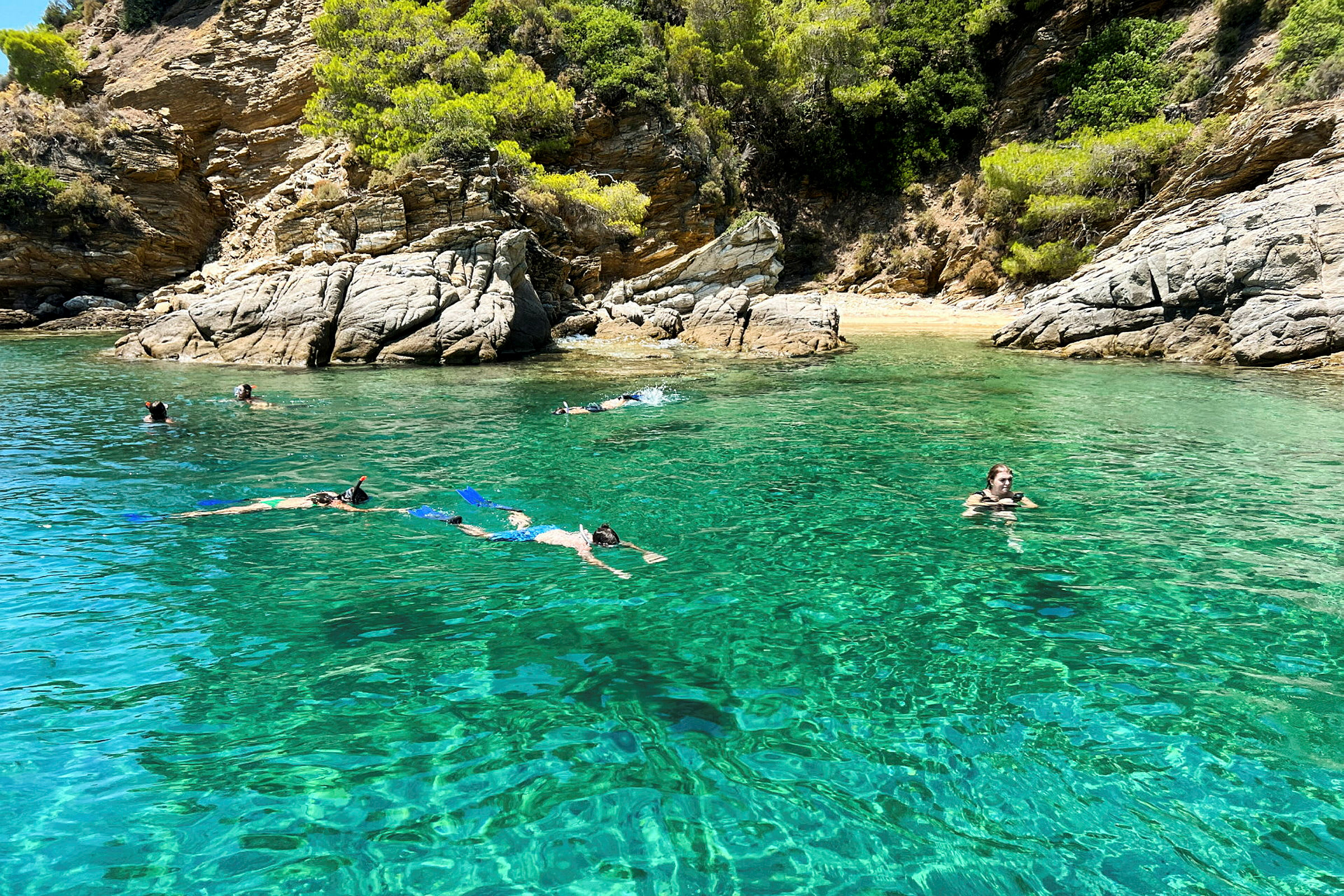 Personer simmar och snorklar i klart turkost vatten nära en klippig strand. (AI-genererad text)