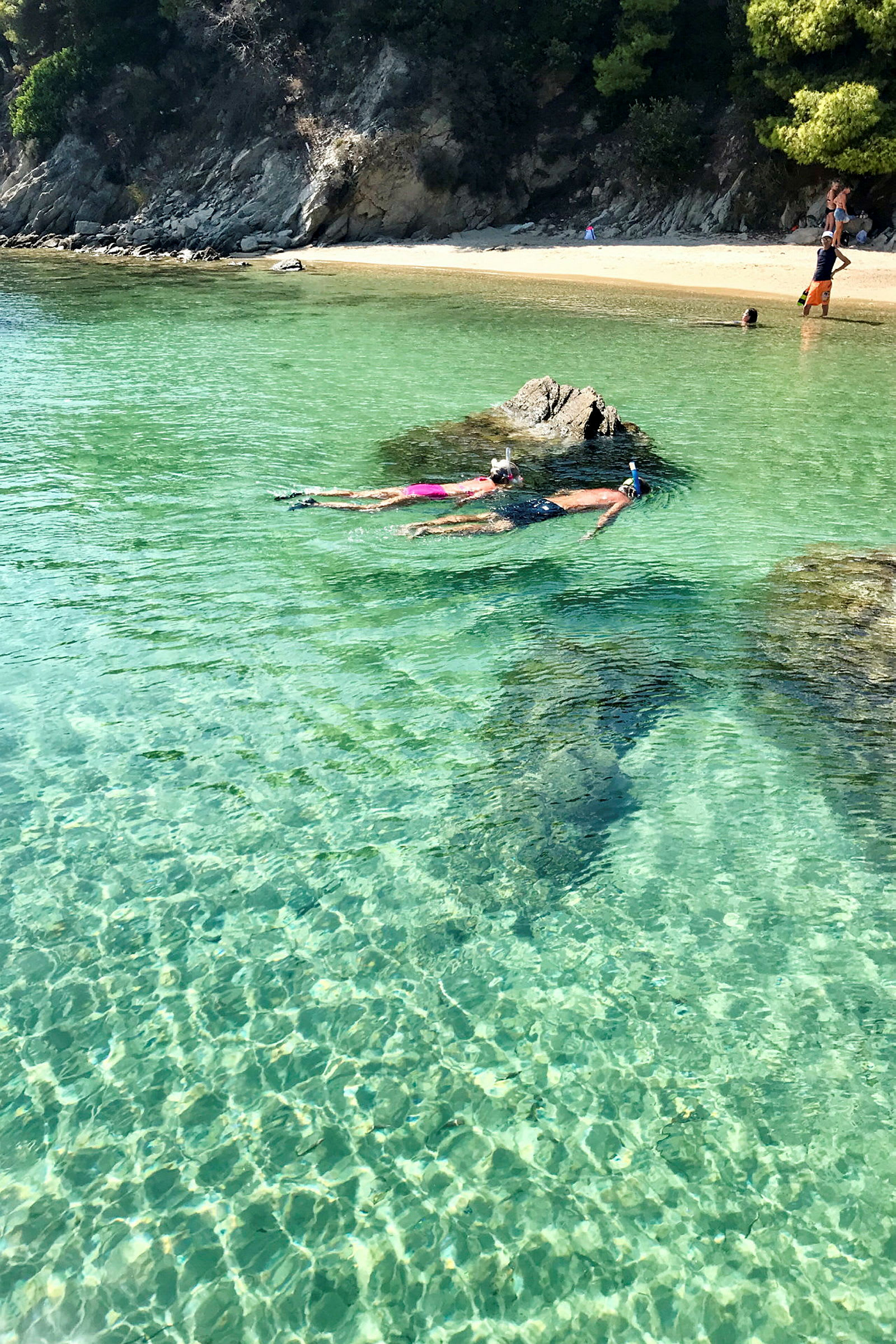 Två personer snorklar i klart, turkost vatten nära en sten, med en sandstrand och träd i bakgrunden. (AI-genererad text)