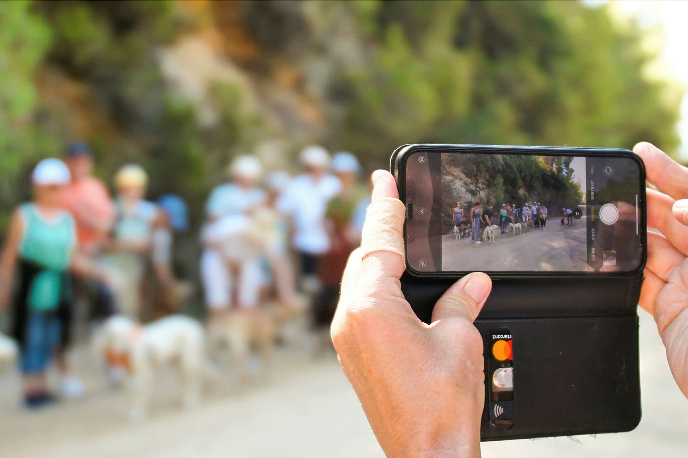 En hand håller en mobiltelefon som tar en bild av en promenadgrupp utomhus. (AI-genererad text)