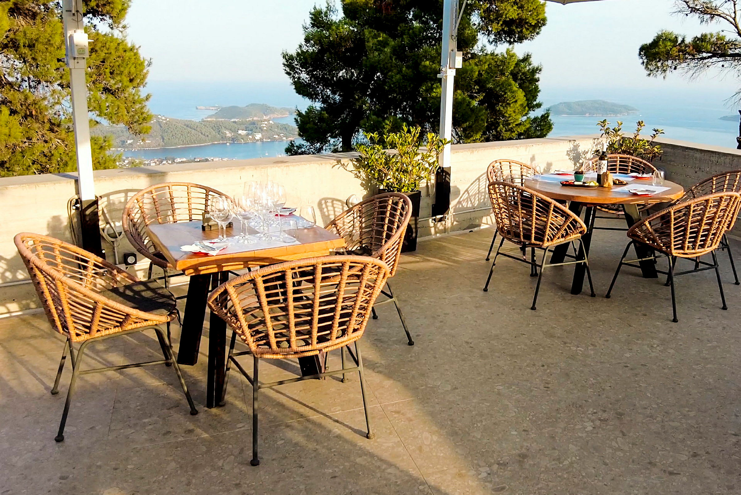 Utomhusrestaurang med bord och stolar i flätad design, med utsikt över havet och grönskande omgivningar. (AI-genererad text)