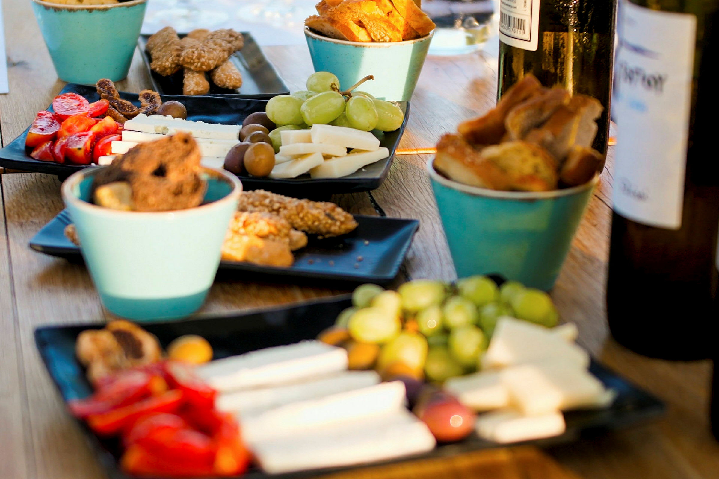 Flera tallrikar innehåller snacks som ost, grönsaker, oliver och bröd, tillsammans med dryckesflaskor på ett bord. (AI-genererad text)