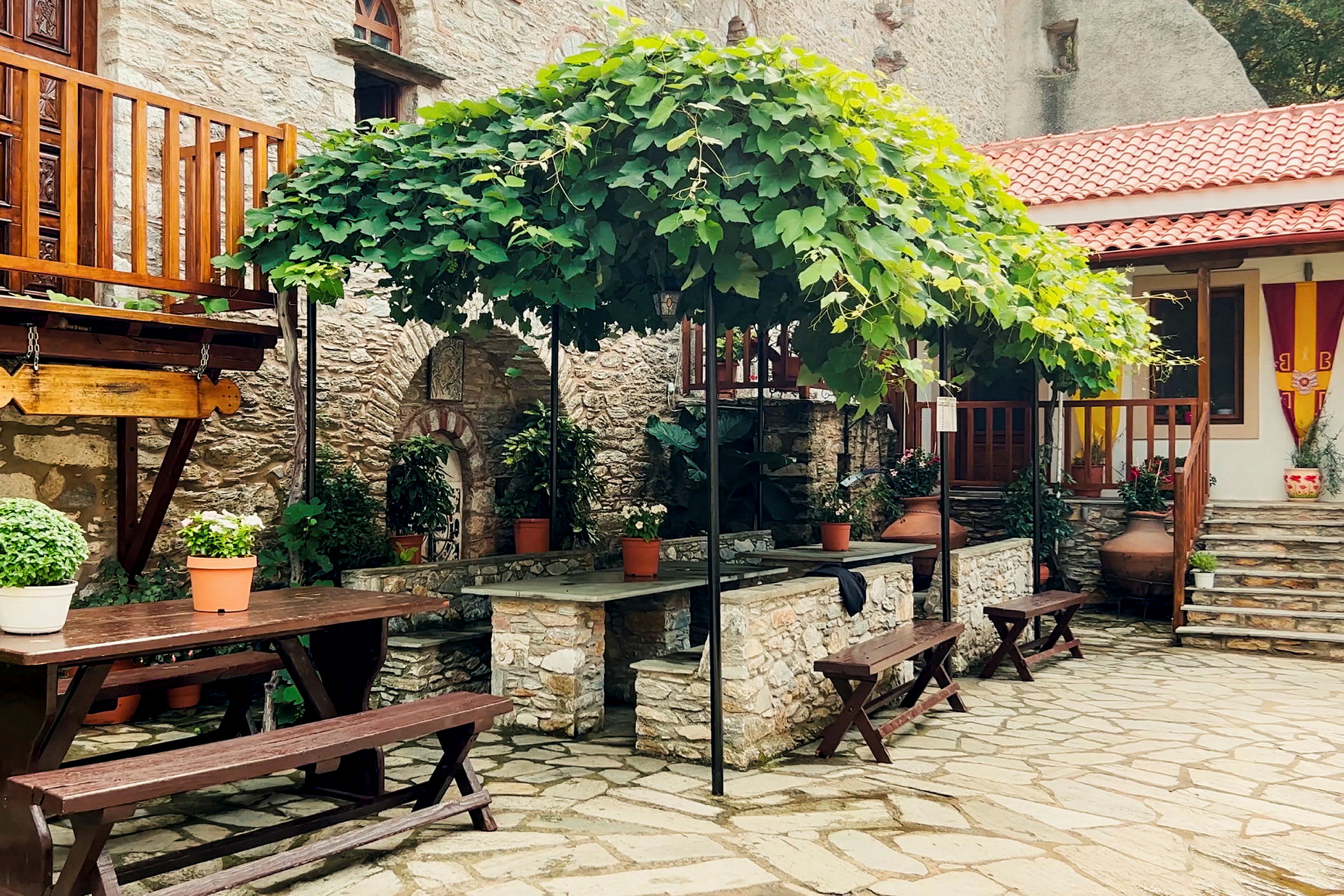 En uteplats med stenlagd mark, träbord och bänkar samt stora blomkrukor. Gröna växter klättrar på en pergola ovanför. (AI-genererad text)
