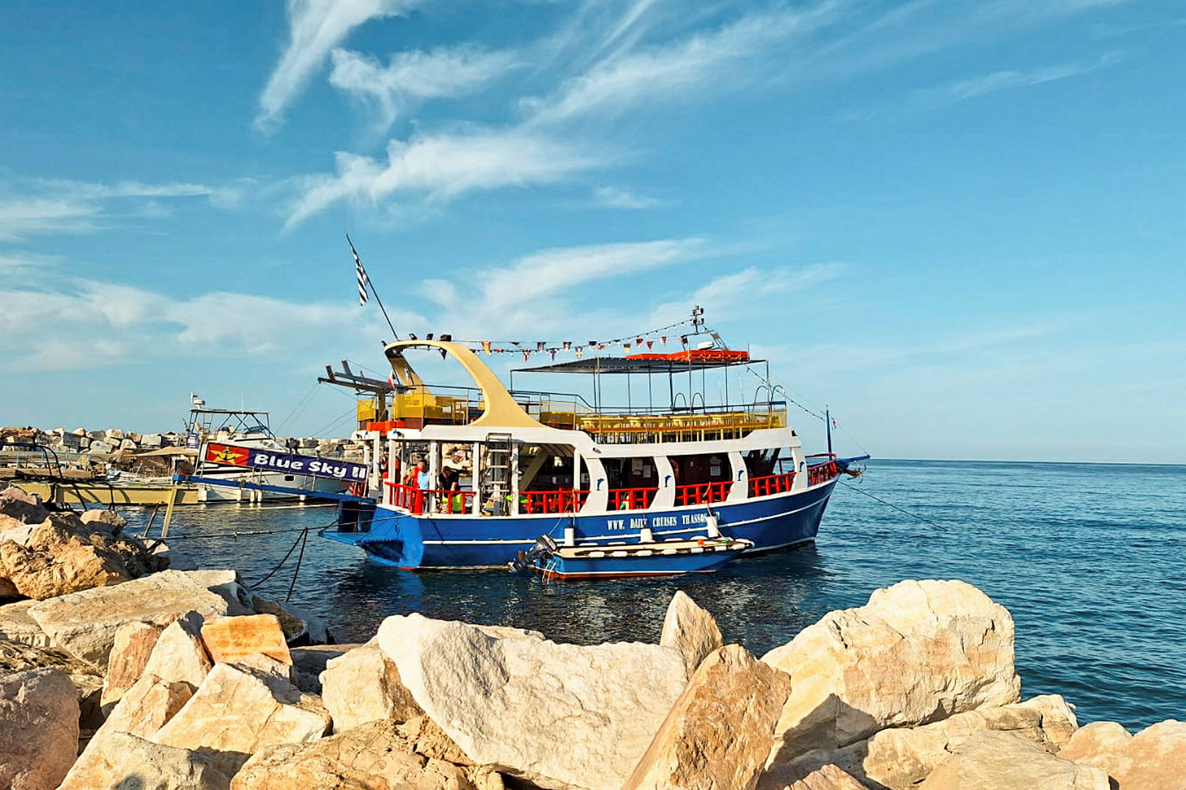 En färgglad turistbåt ligger vid kajen nära stora, ljusa stenar. Havet och en klar himmel syns i bakgrunden. (AI-genererad text)