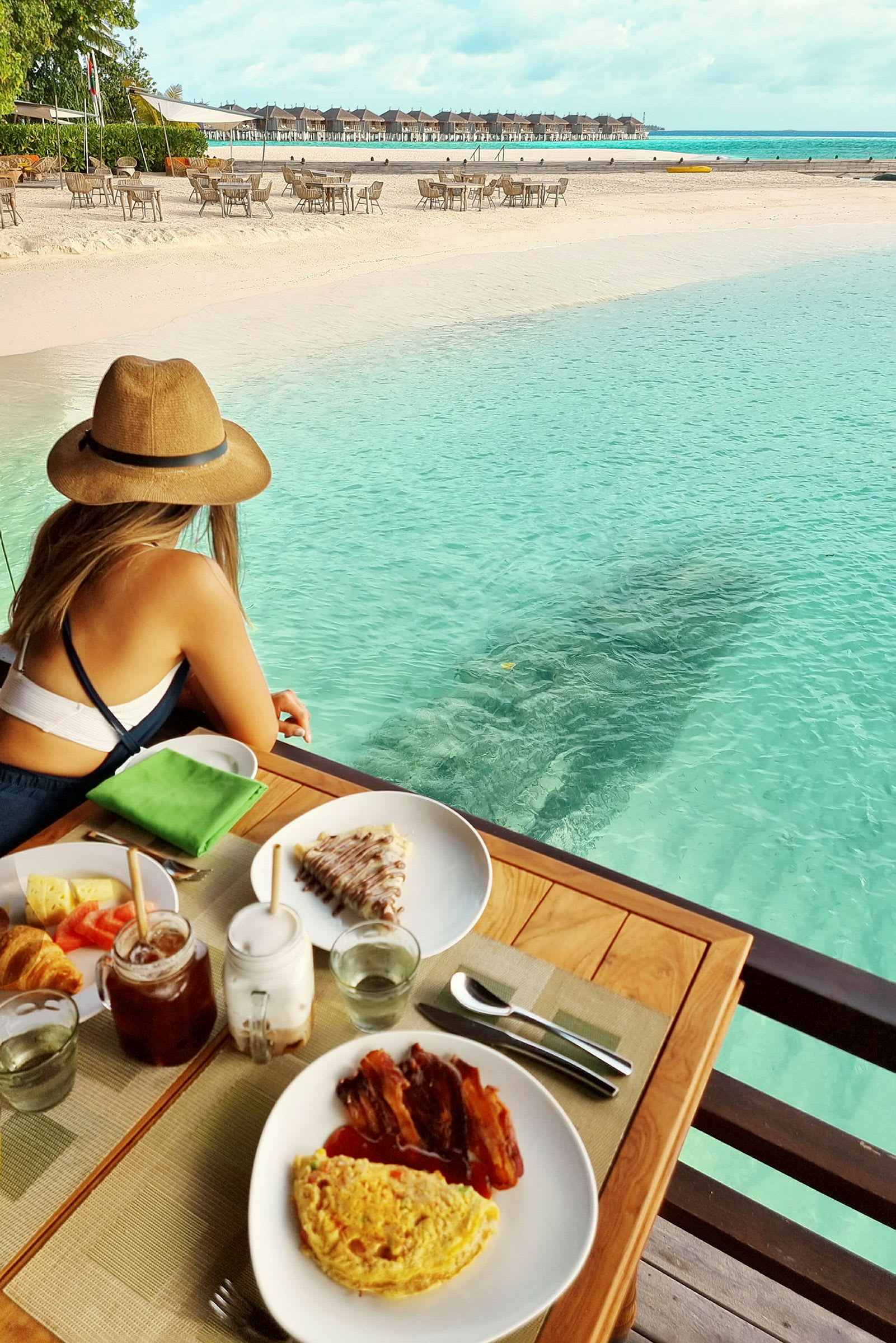 En kvinna sitter vid ett bord med frukostmat, vid strandkanten med turkost vatten och utsikt över strand och bungalows. (AI-genererad text)