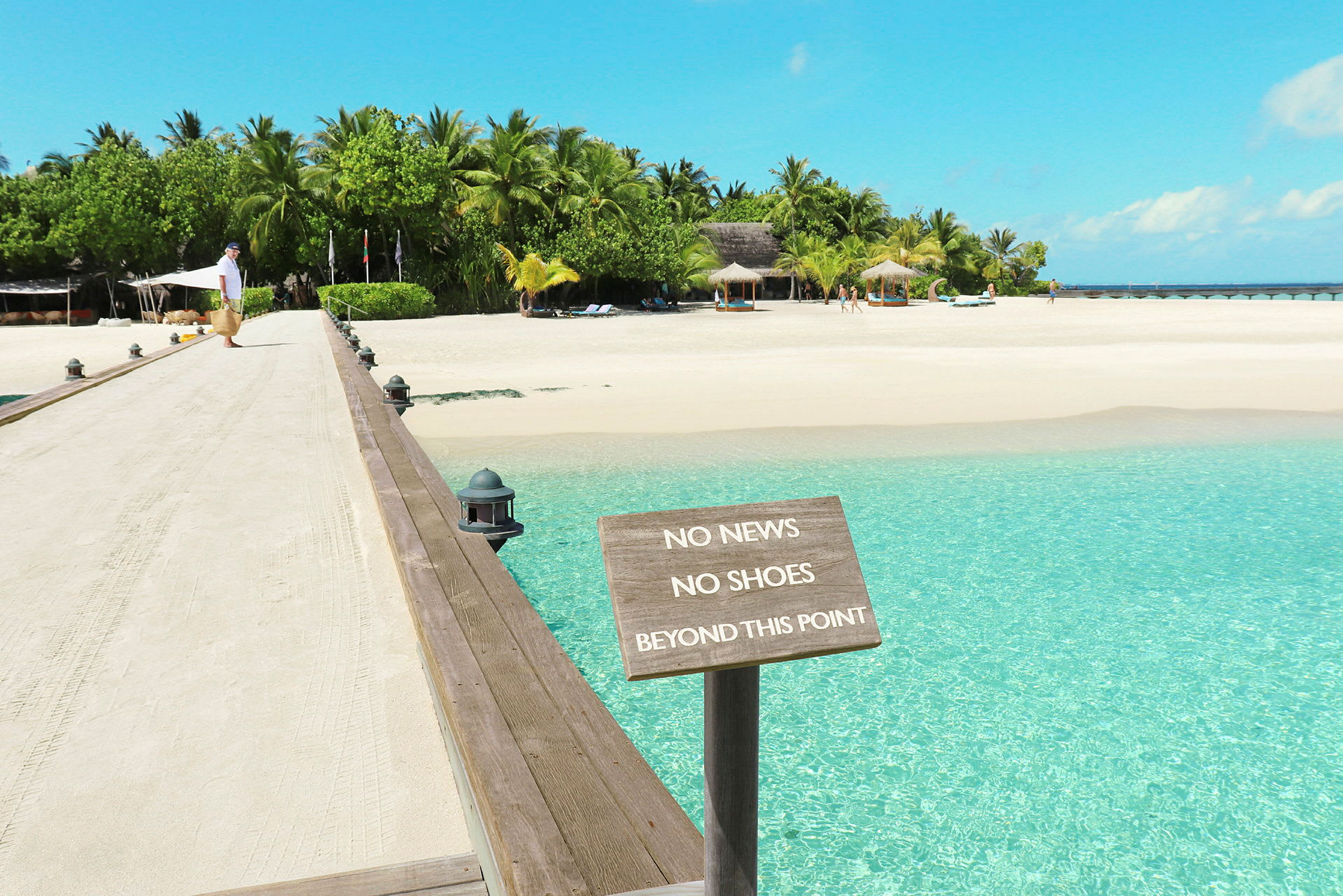 En sandig gångbro leder till en tropisk strand med en skylt som har texten 'No News No Shoes Beyond This Point'. (AI-genererad text)