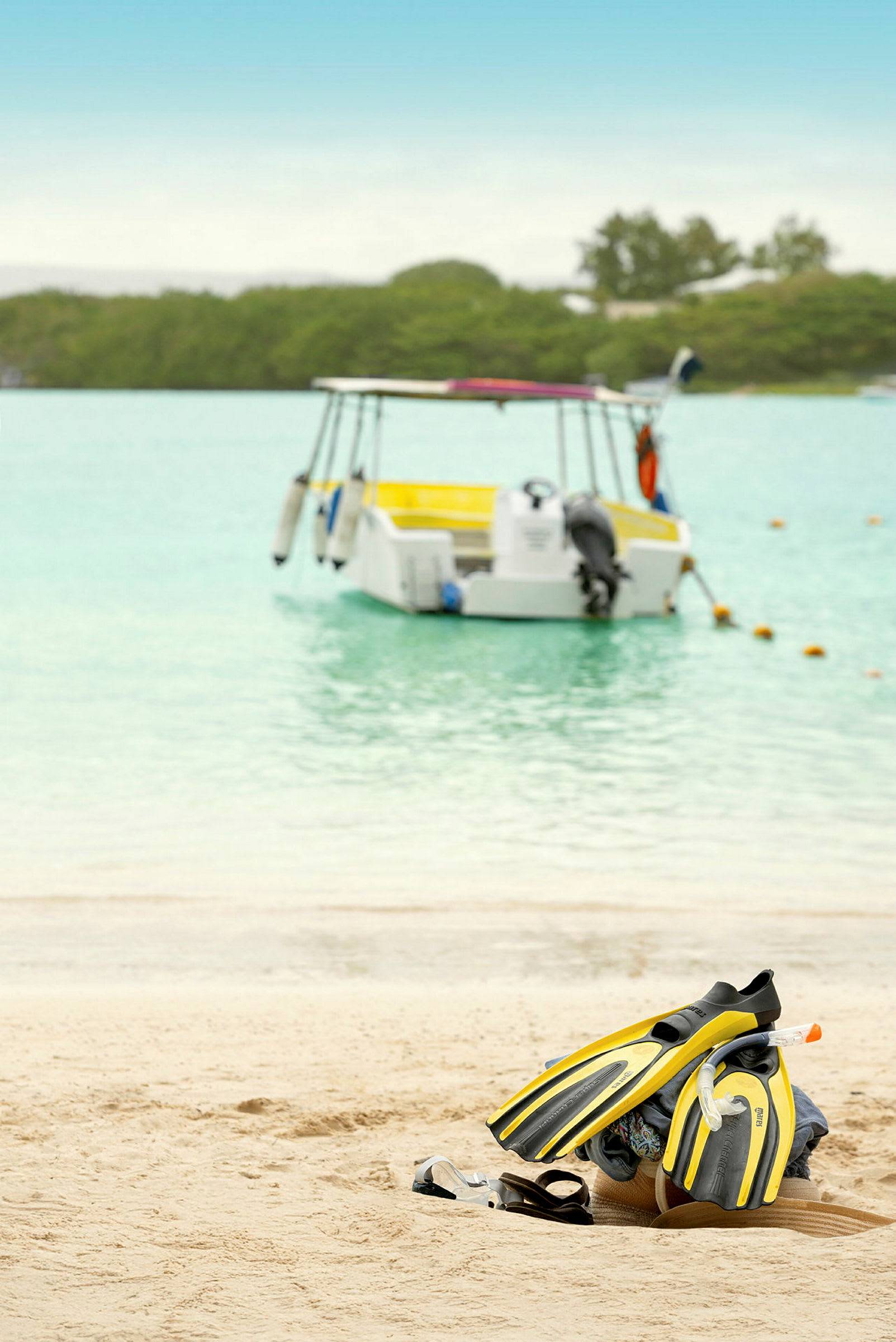 Ett par gulsvarta simfenor, snorkelutrustning och sandaler är placerade på en sandstrand. En båt är förankrad i vattnet i bakgrunden. (AI-genererad text)