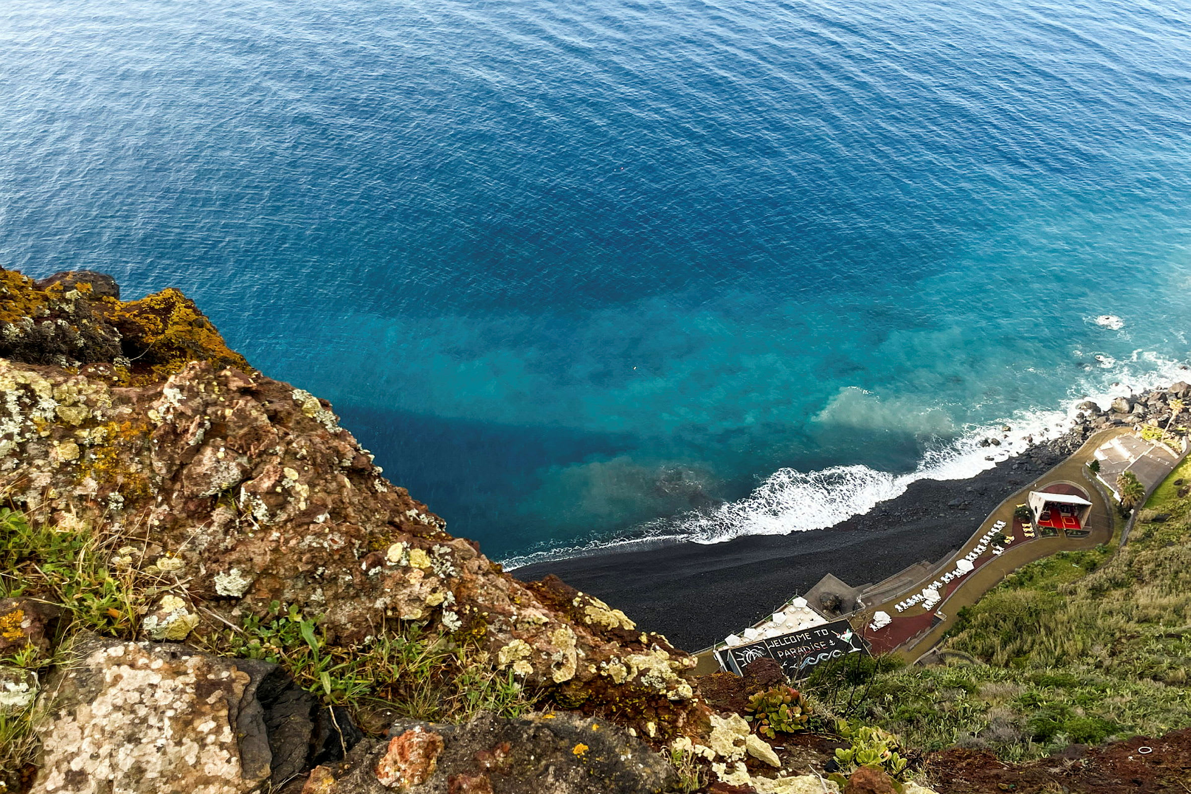 Utsikt från en klippa ned mot en kustlinje med stenstrand och byggnader, samt havet. (AI-genererad text)
