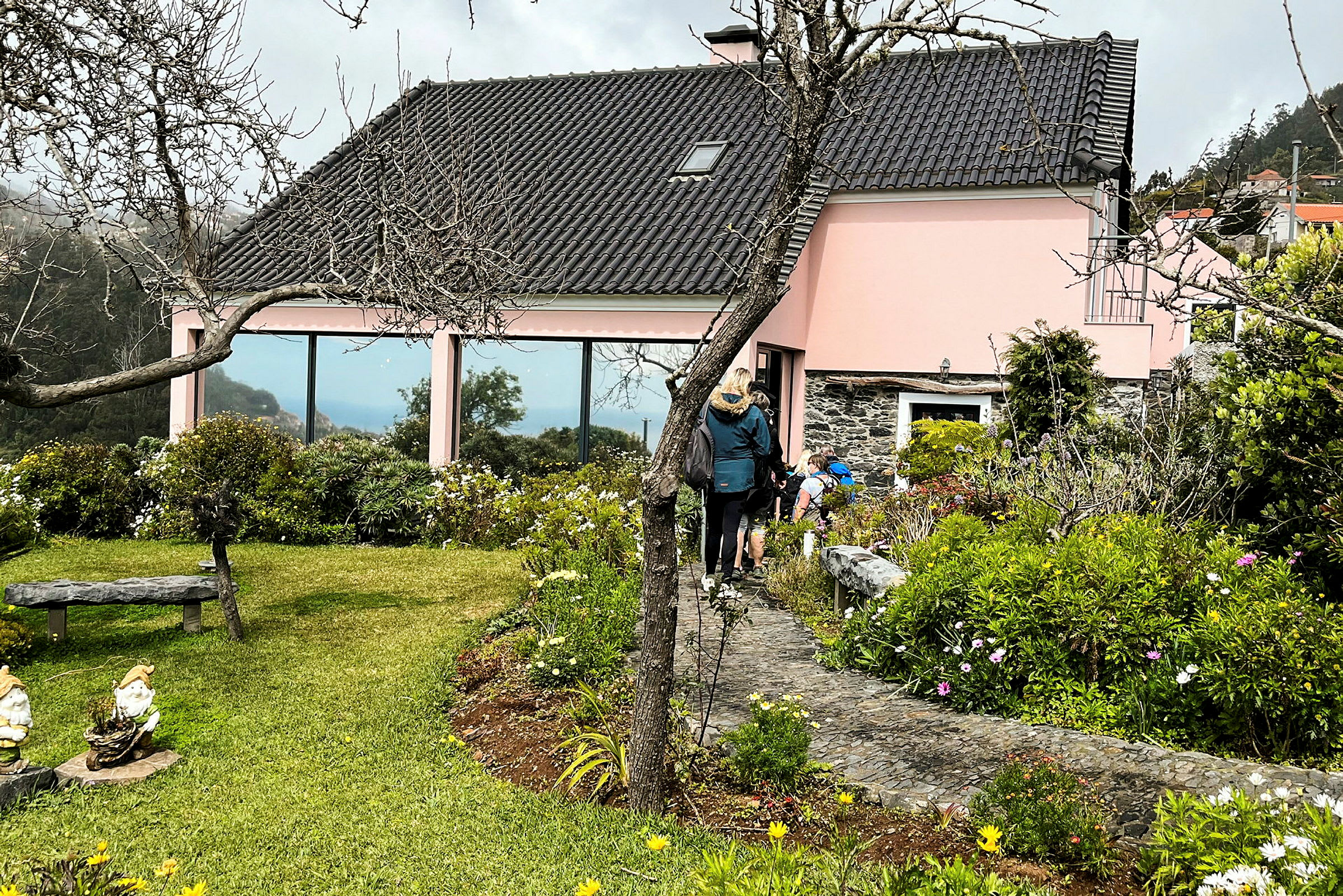 En rosa byggnad med svart tak omgiven av grönska och blommor, med personer som går på en gångväg. (AI-genererad text)