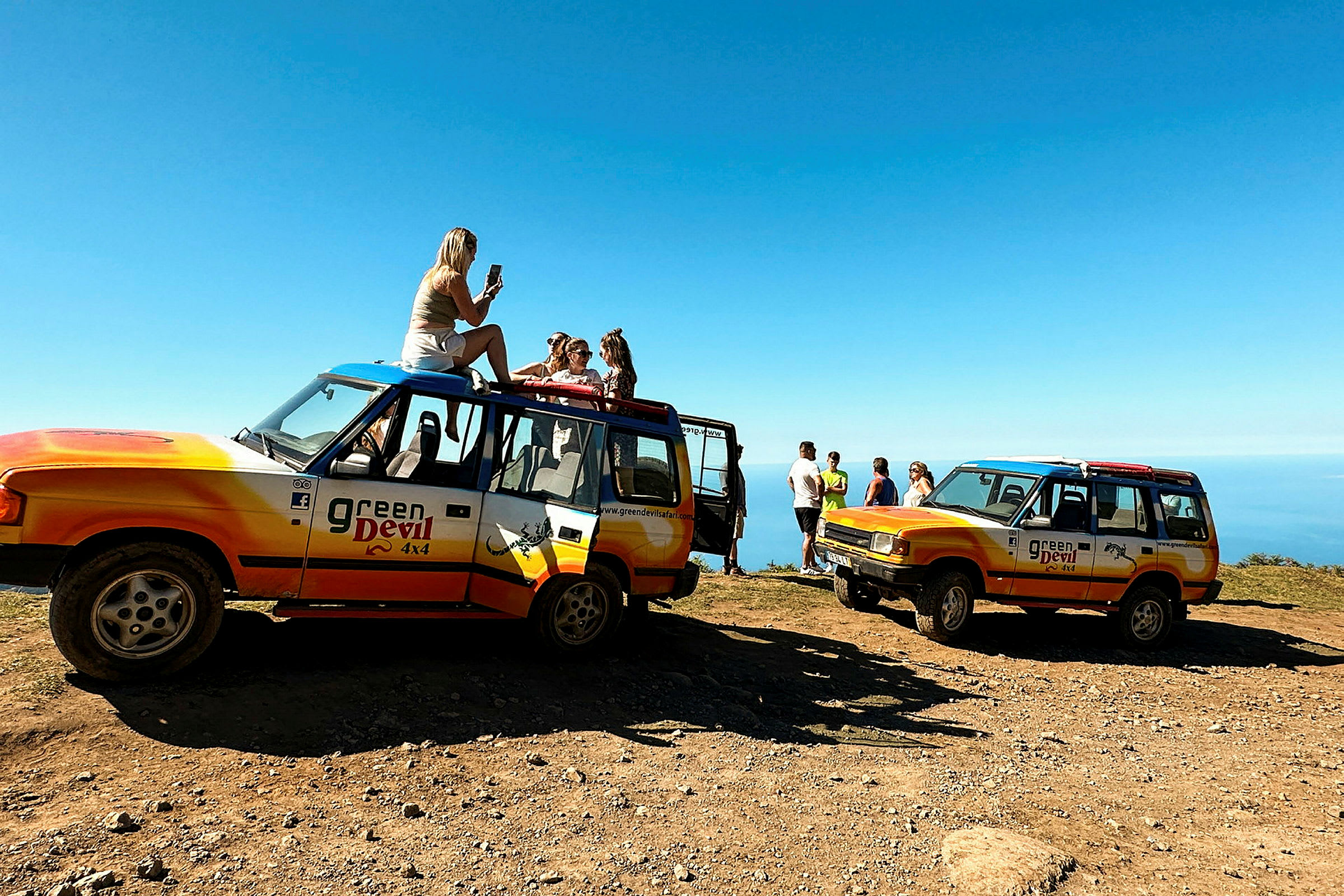 Två offroadfordon med logotypen "Green Devil 4x4" är parkerade på en grusväg med utsikt över havet. Människor står eller sitter nära fordonen. (AI-genererad text)