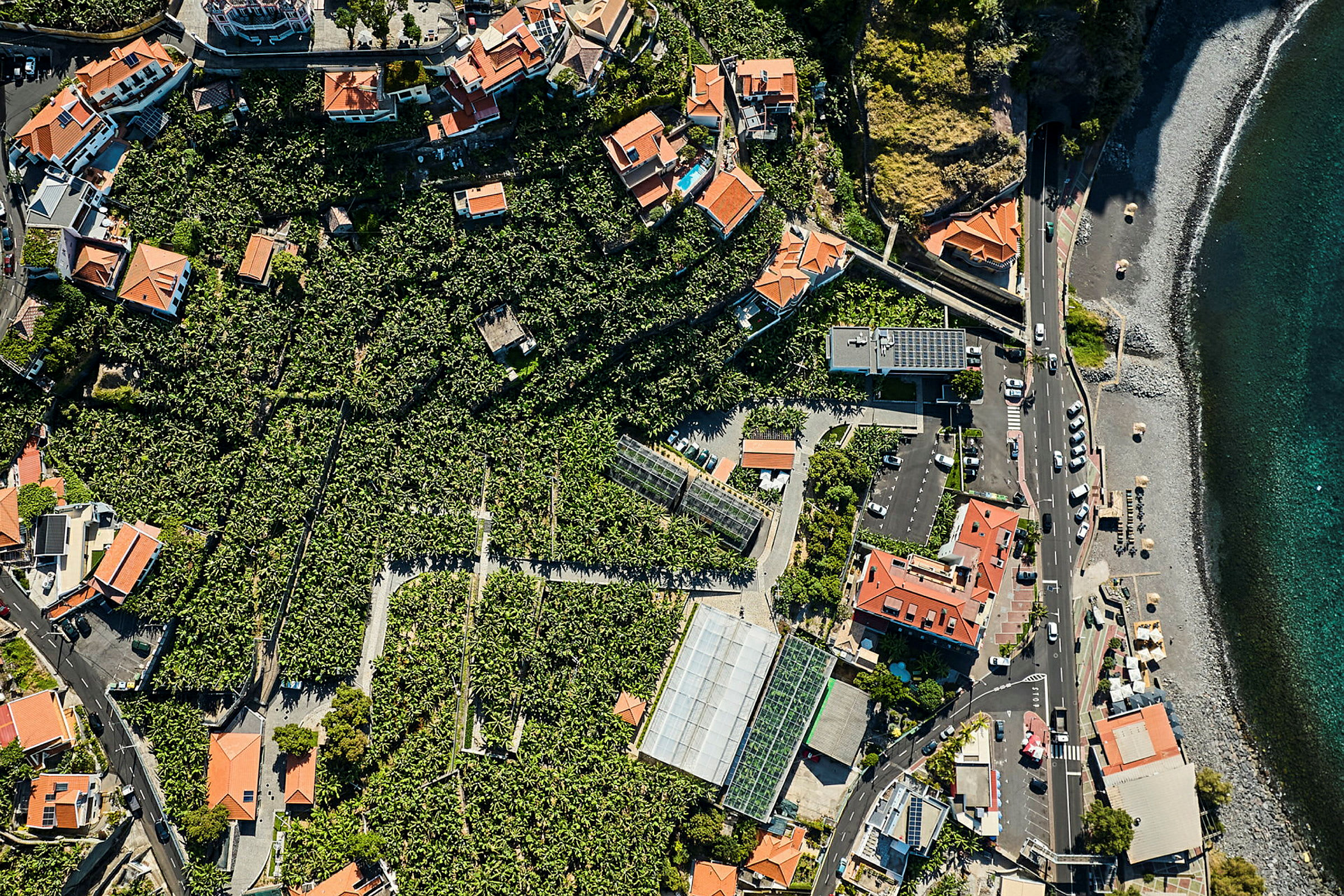 Flygfoto över ett område med hus med röda tak, omgivna av grön vegetation och nära en strand med flera bilar parkerade längs en väg. (AI-genererad text)