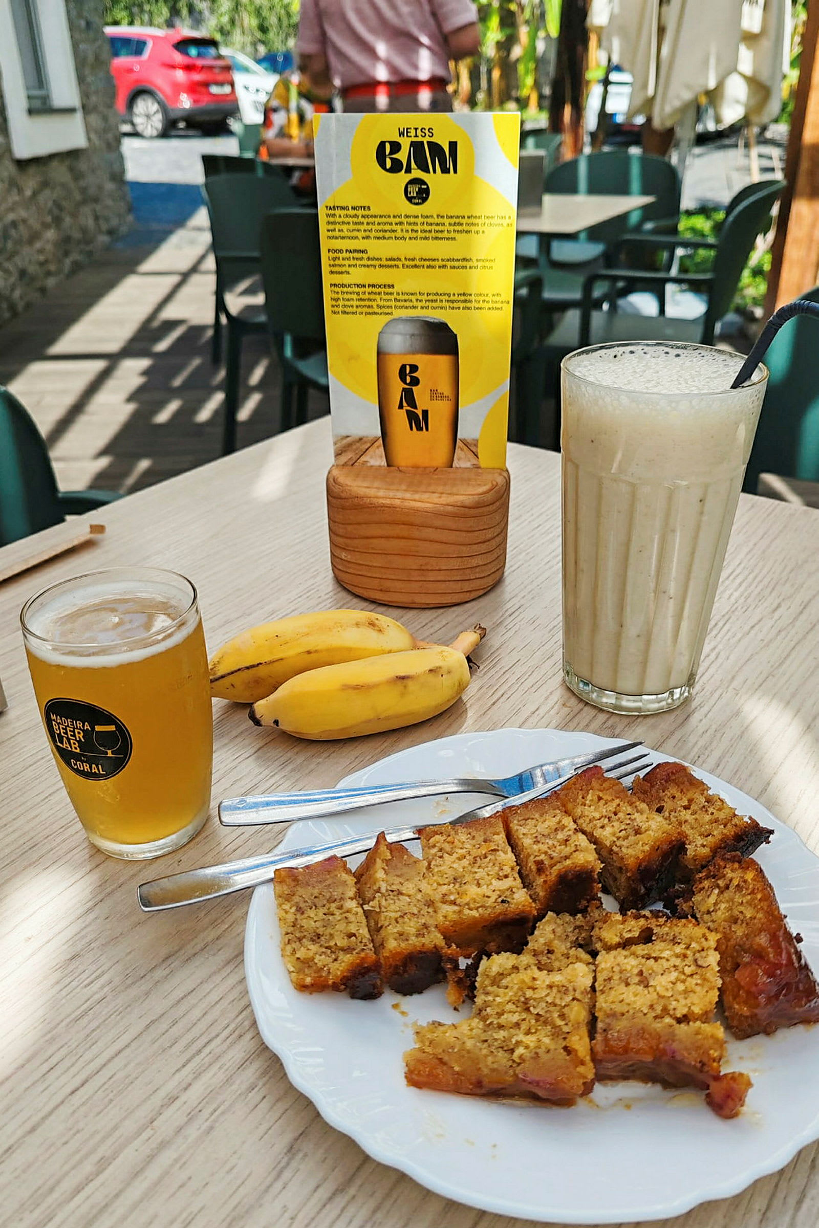 En tallrik med skivor av kaka, två bananer, ett glas öl och en milkshake på ett bord. Bakom syns ett menykort och sittmöbler utomhus. (AI-genererad text)