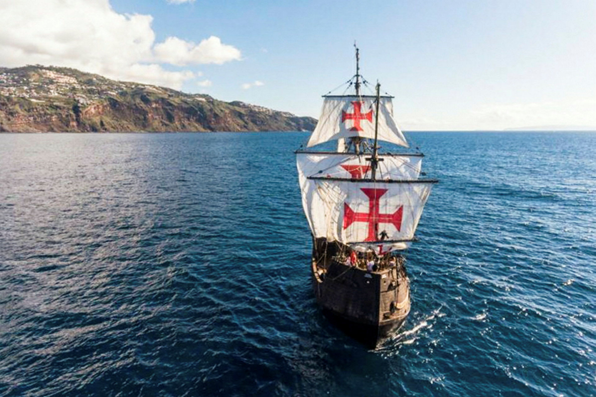 Ett segelfartyg med stora vita segel dekorerade med röda kors navigerar över en blå ocean i närheten av en kuperad kust. (AI-genererad text)