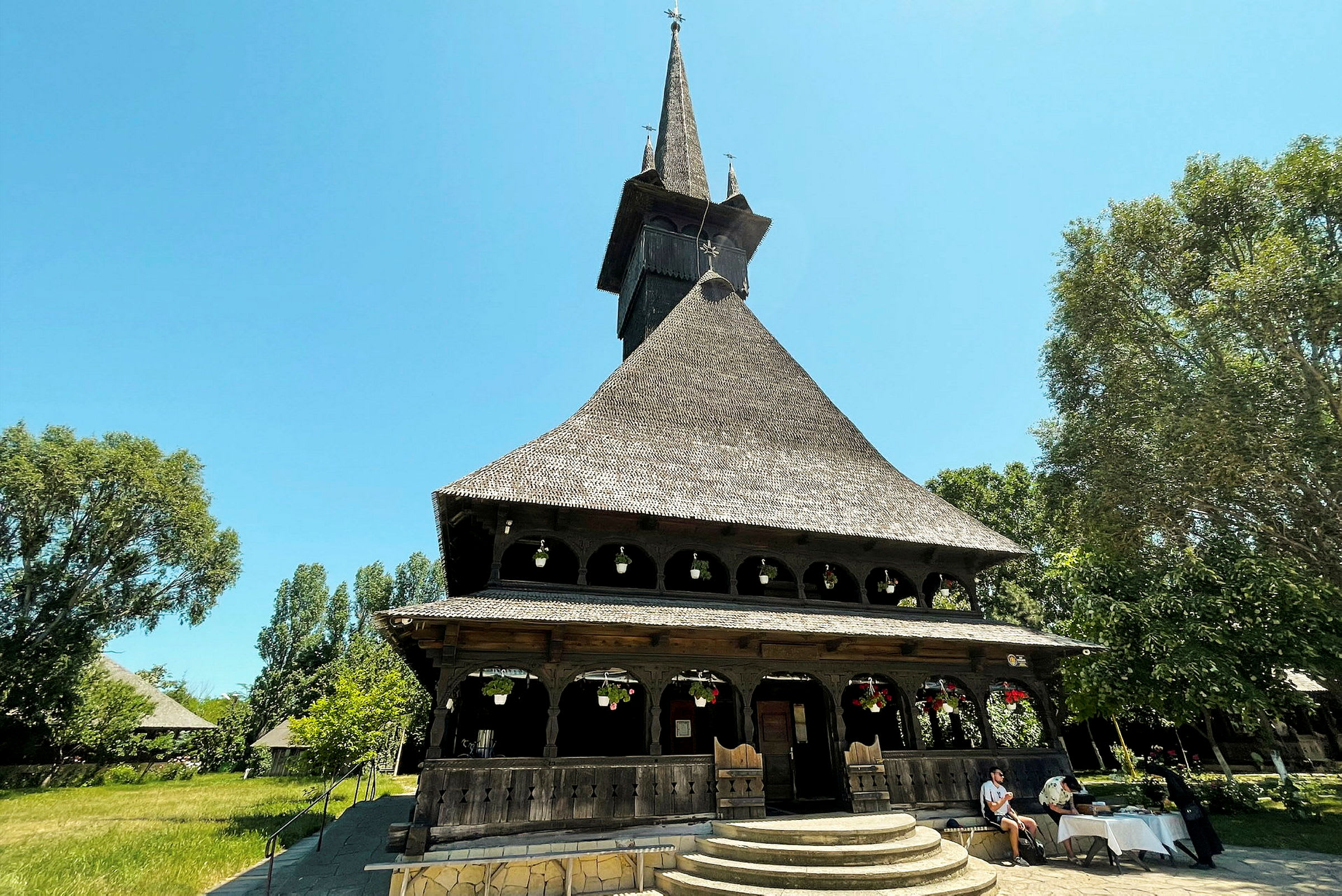 Närbild av en träkyrka med spetsigt tak och dekorativa element, omgiven av grönska och människor. (AI-genererad text)
