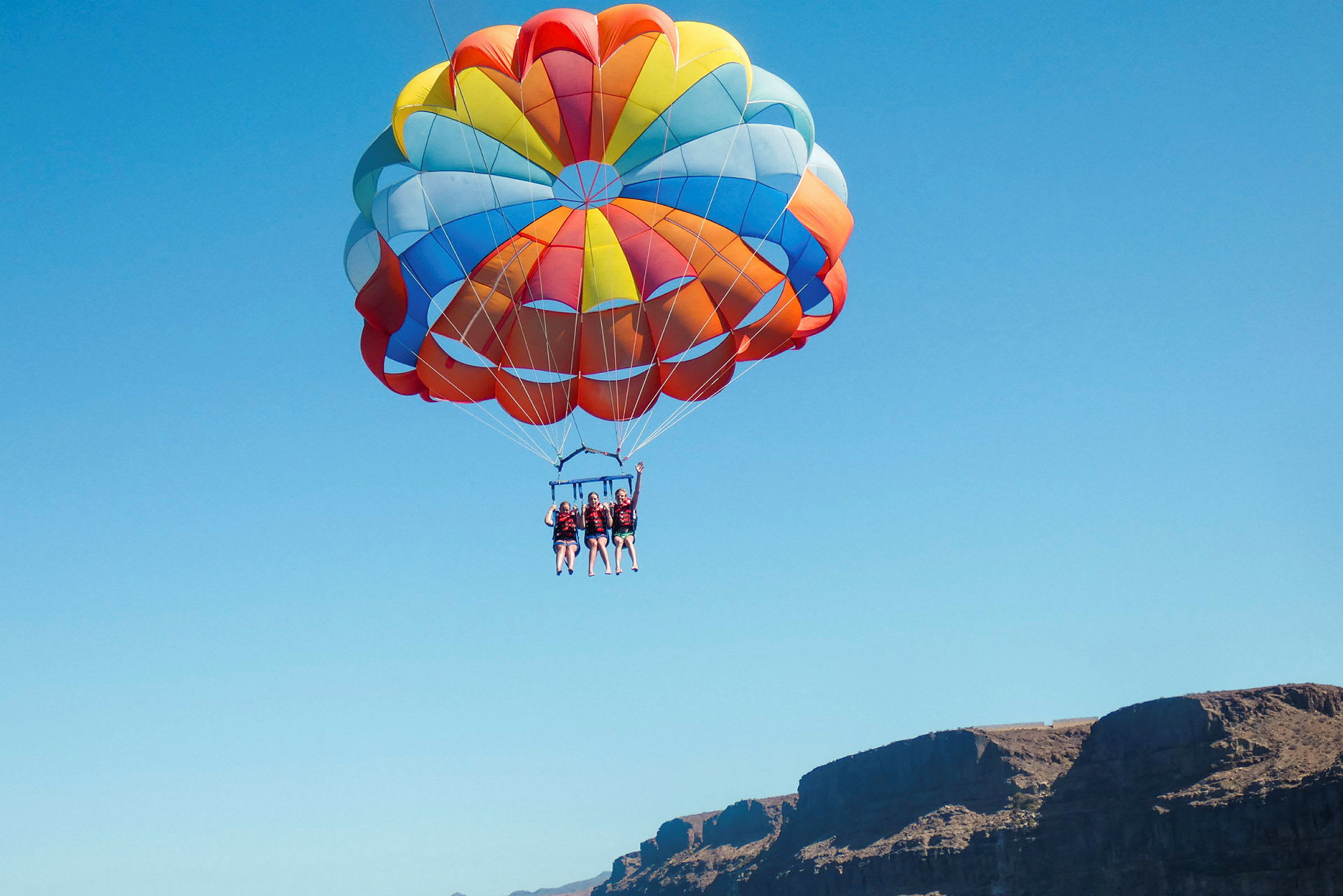 Tre personer som parasailar med en stor, färgglad fallskärm mot en blå himmel. (AI-genererad text)