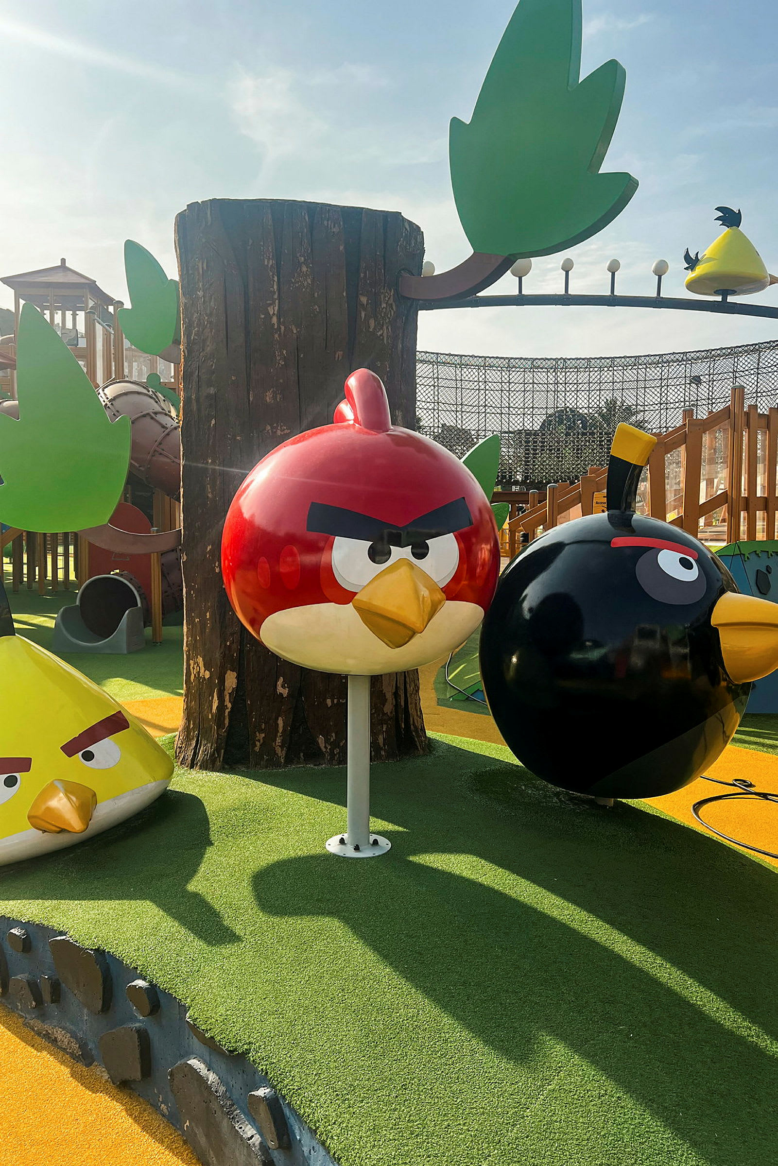Lekplats med Angry Birds-figurer, inklusive en röd fågel, en svart fågel och en gul trekantig fågel, med träd och andra lekstrukturer runtomkring. (AI-genererad text)