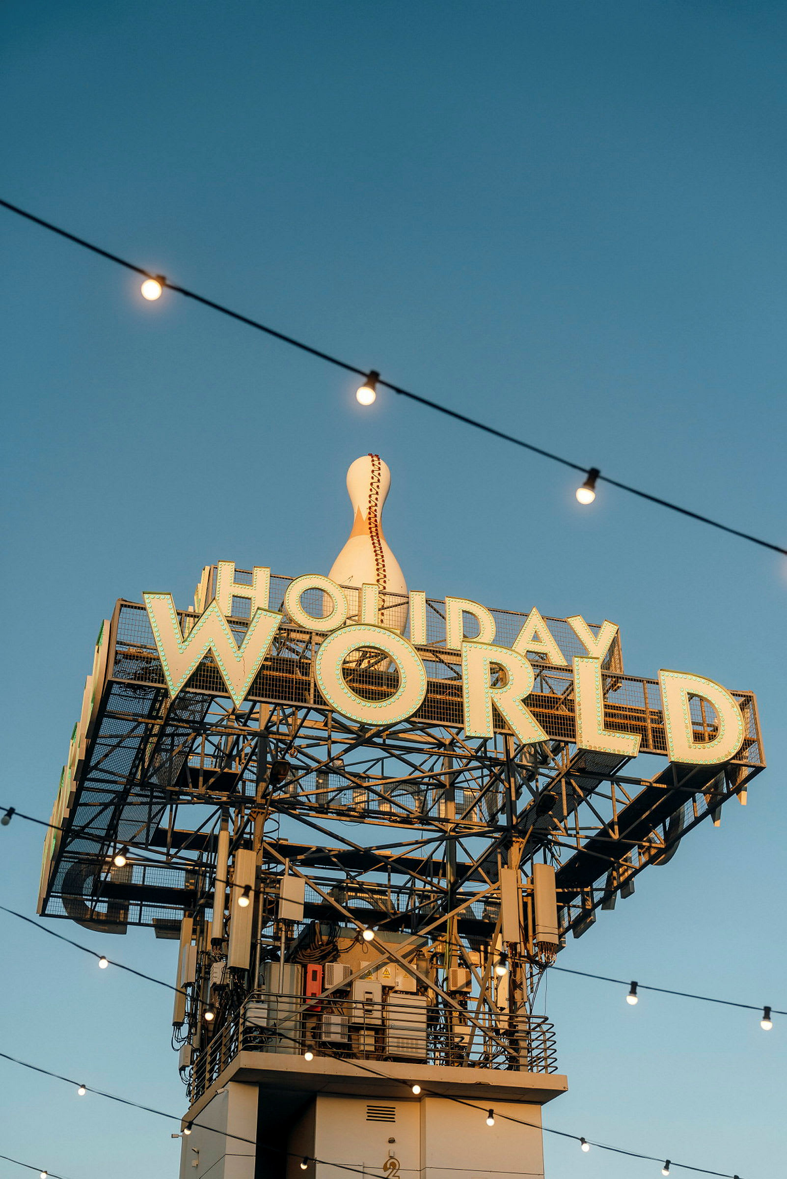 Skylt med texten 'Holiday World' och en stor dekorerad bowlingkägla ovanpå, med ljusslingor mot en klarblå himmel. (AI-genererad text)